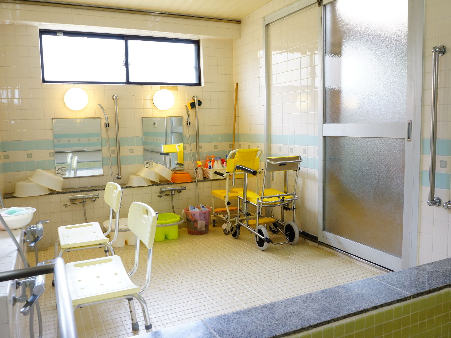 白を基調にした明るい雰囲気の浴室は随所に手摺りを設置。椅子やたらい、車椅子などのほかにスリッパ等もおかれている。