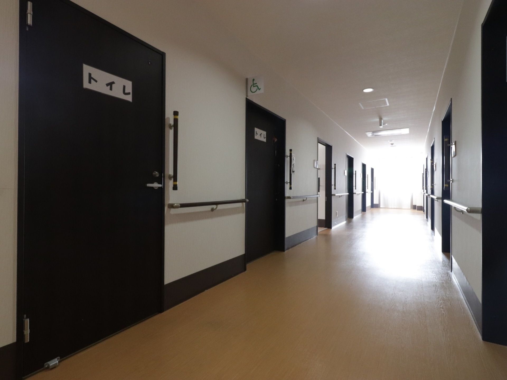 トイレ前の廊下の様子。まっすぐ続いている廊下には手すりがあり、ドアが並んでいる。突き当たりには、窓がある。床はフローリングである。