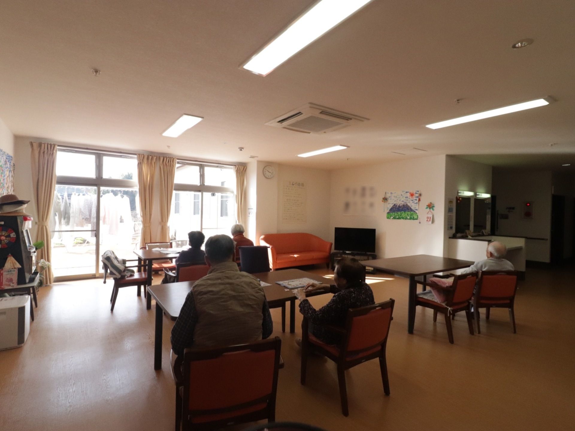 リビングの様子。テーブルが3つ置かれていて、入居者様たちが椅子に座っている。奥にテレビが設置されている。