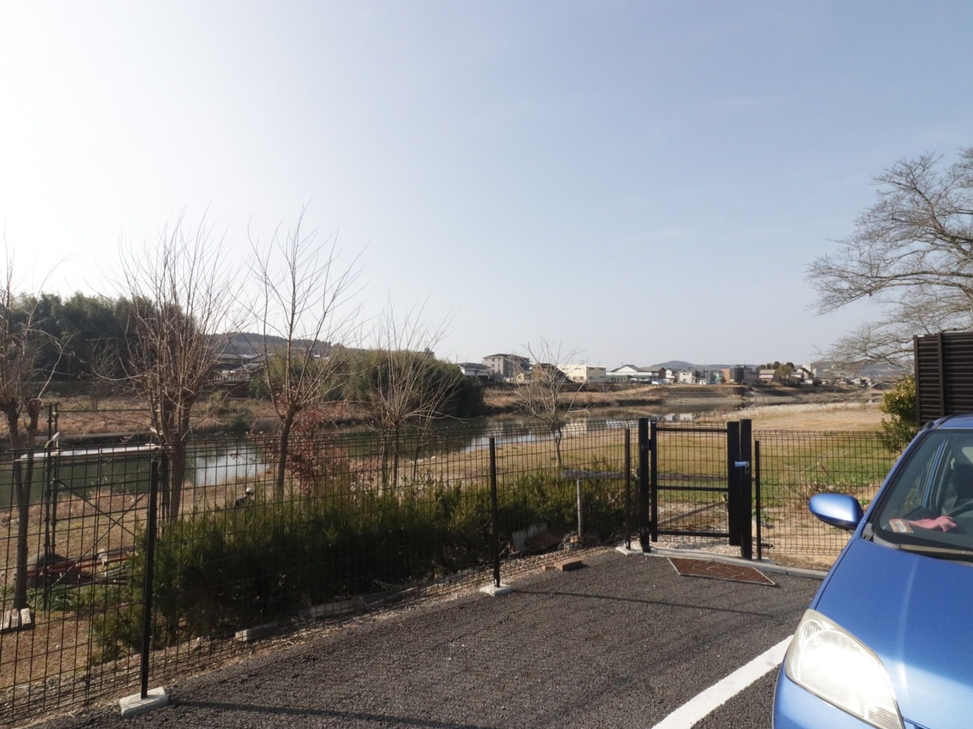 手前に施設の駐車スペースがあり、車が停まっている。フェンスにドアが付いており、河川敷に出られるようになっている。