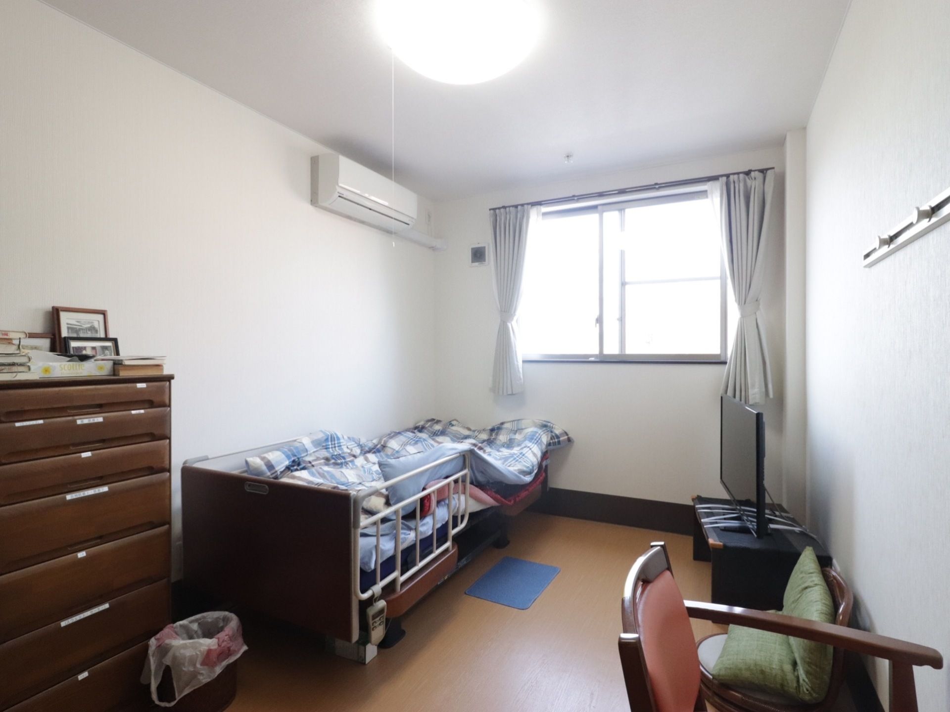 ある入居者様の居室。奥に窓があり、カーテンが付いている。エアコンが完備され、ベッドやタンス、テレビが置かれている。
