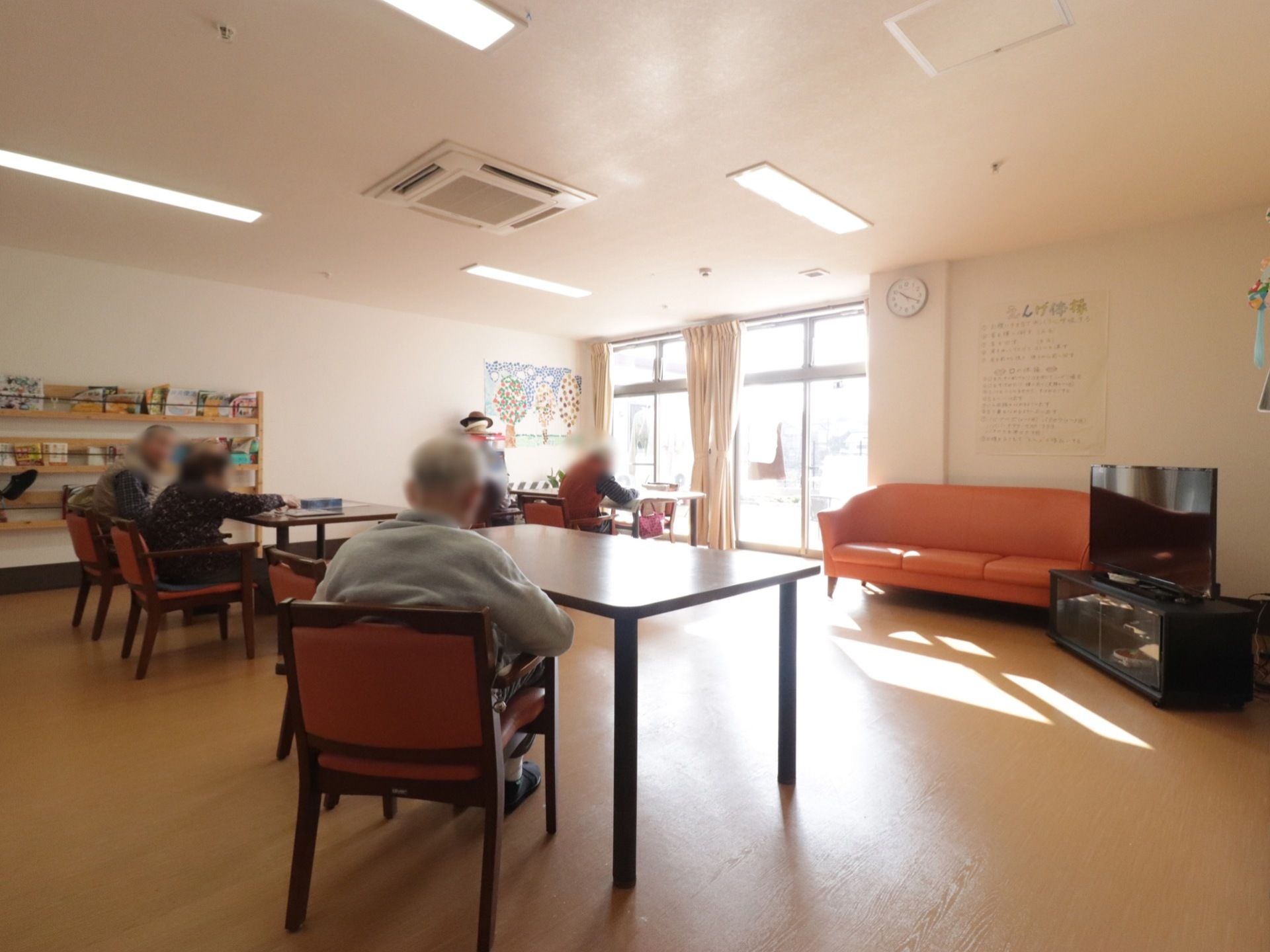 リビングの奥に大きな窓がある。左奥の壁に手作りの作品が展示され、本などが並べられているラックが設置されている。