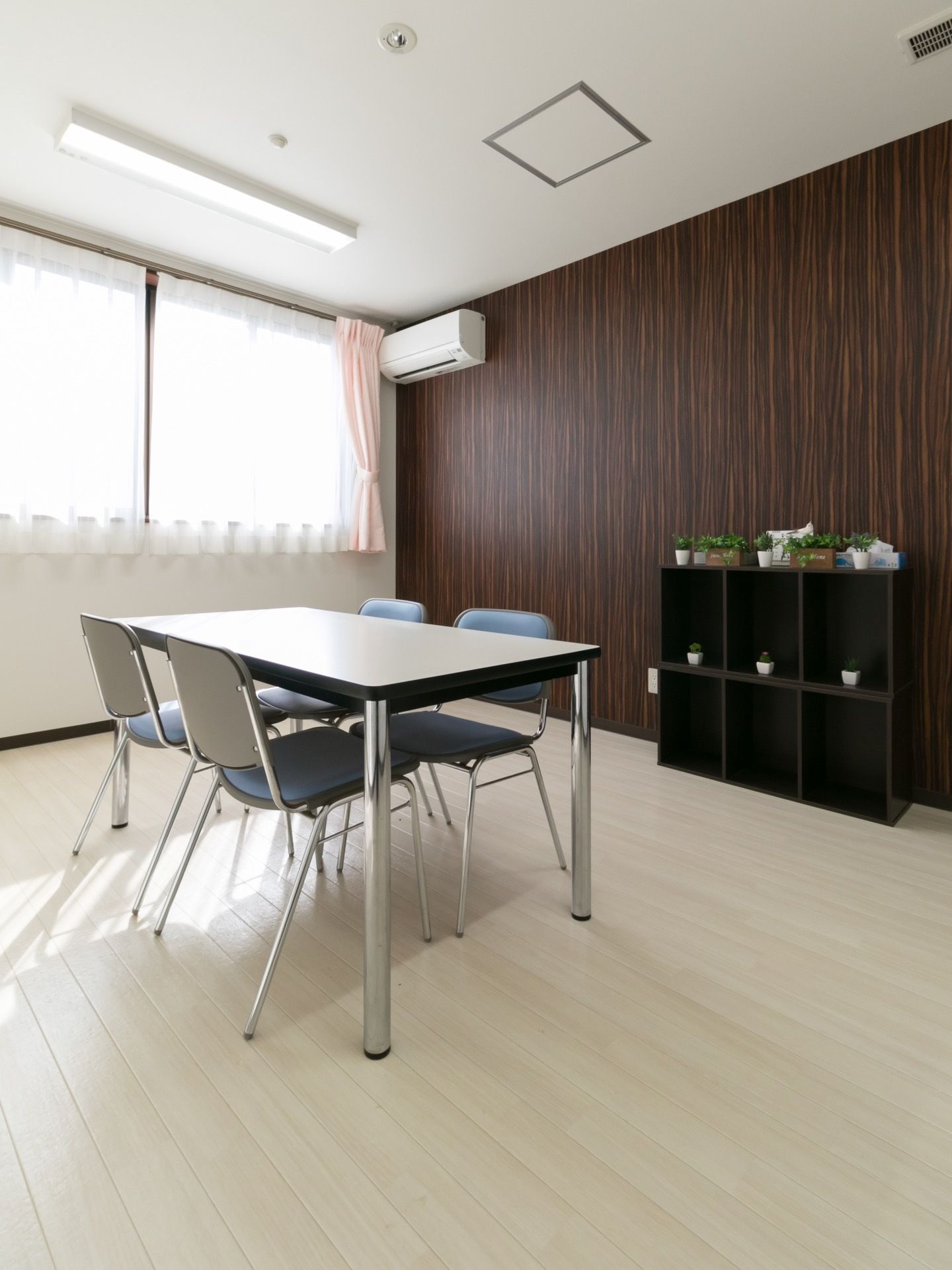 テーブルと椅子を配備し、白い木目調を床に、こげ茶を壁の一面だけに利用している。壁面には、こげ茶のカラーボックスの上に緑の植物が飾られている。