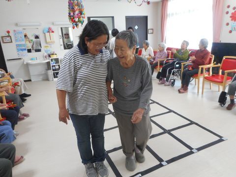 床に黒い枠のようなものが置いてあり、そこを歩く高齢女性を介助する女性職員