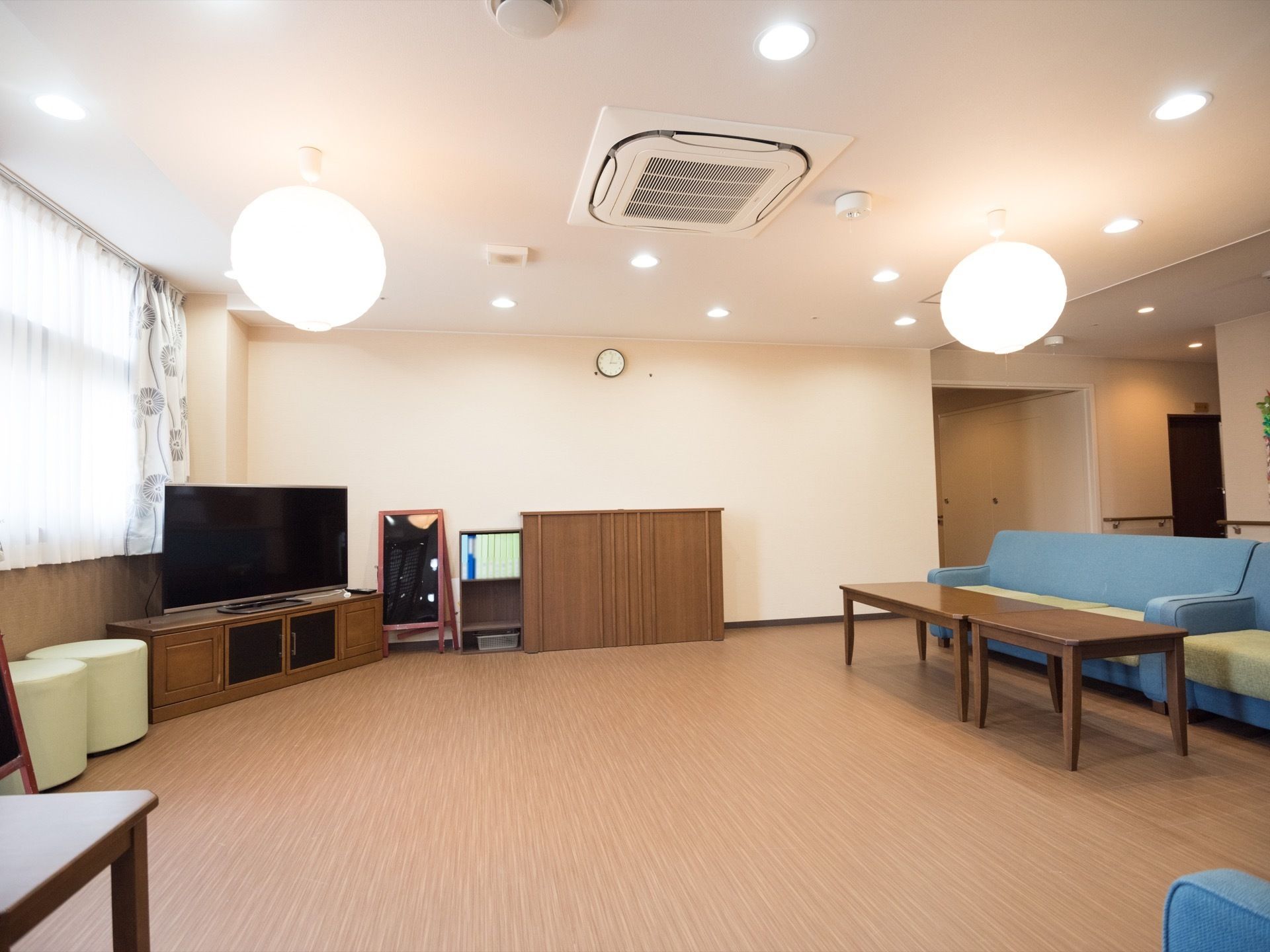 窓の手前に茶色いテレビ台があり、大画面のテレビが乗せられている。テレビを囲むように青い大きなソファが複数設置されている。