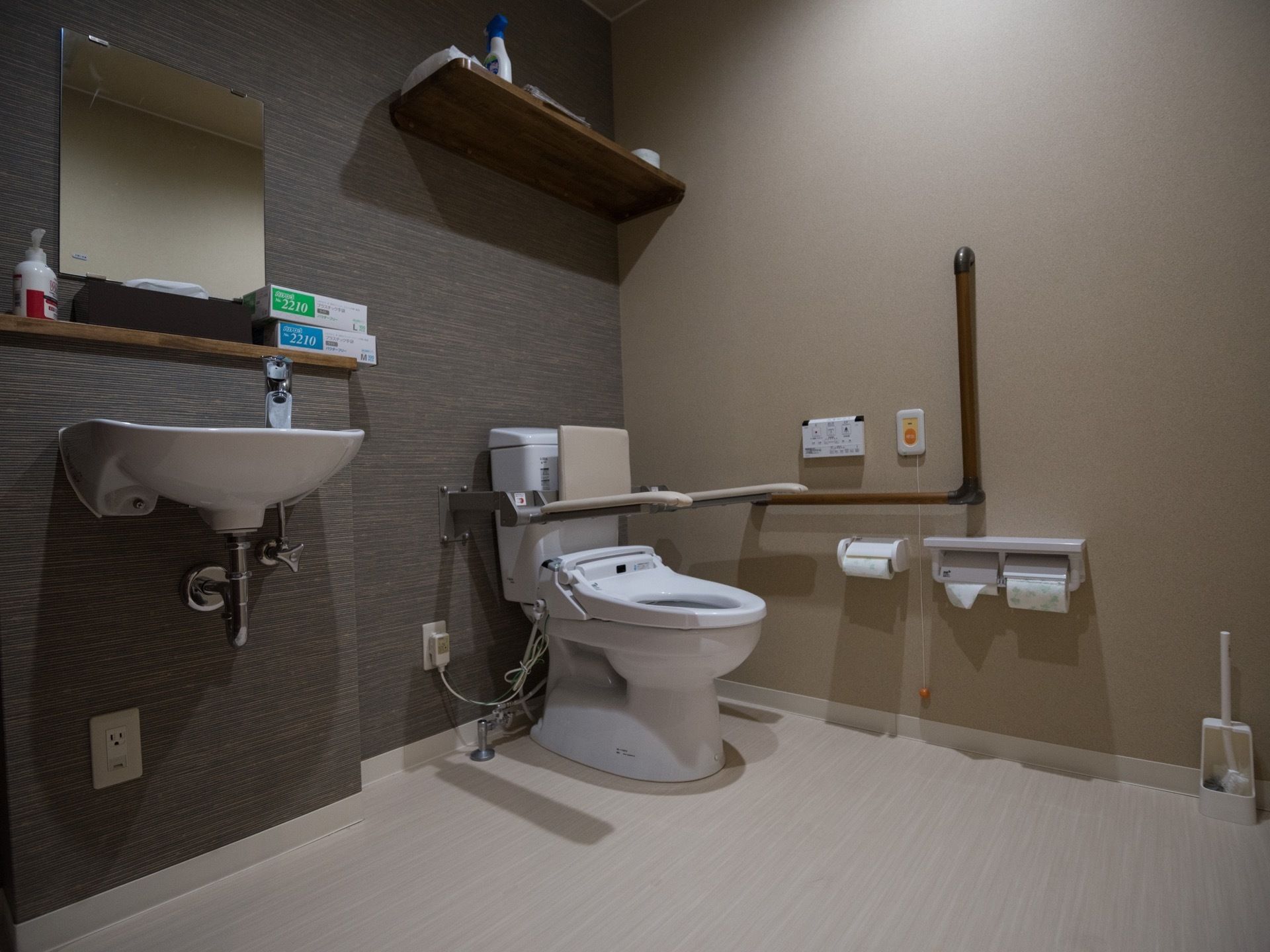 トイレには肘掛けと背もたれが取りつけられている。横の壁にはＬ字型の手すりと緊急コールが設置されている。