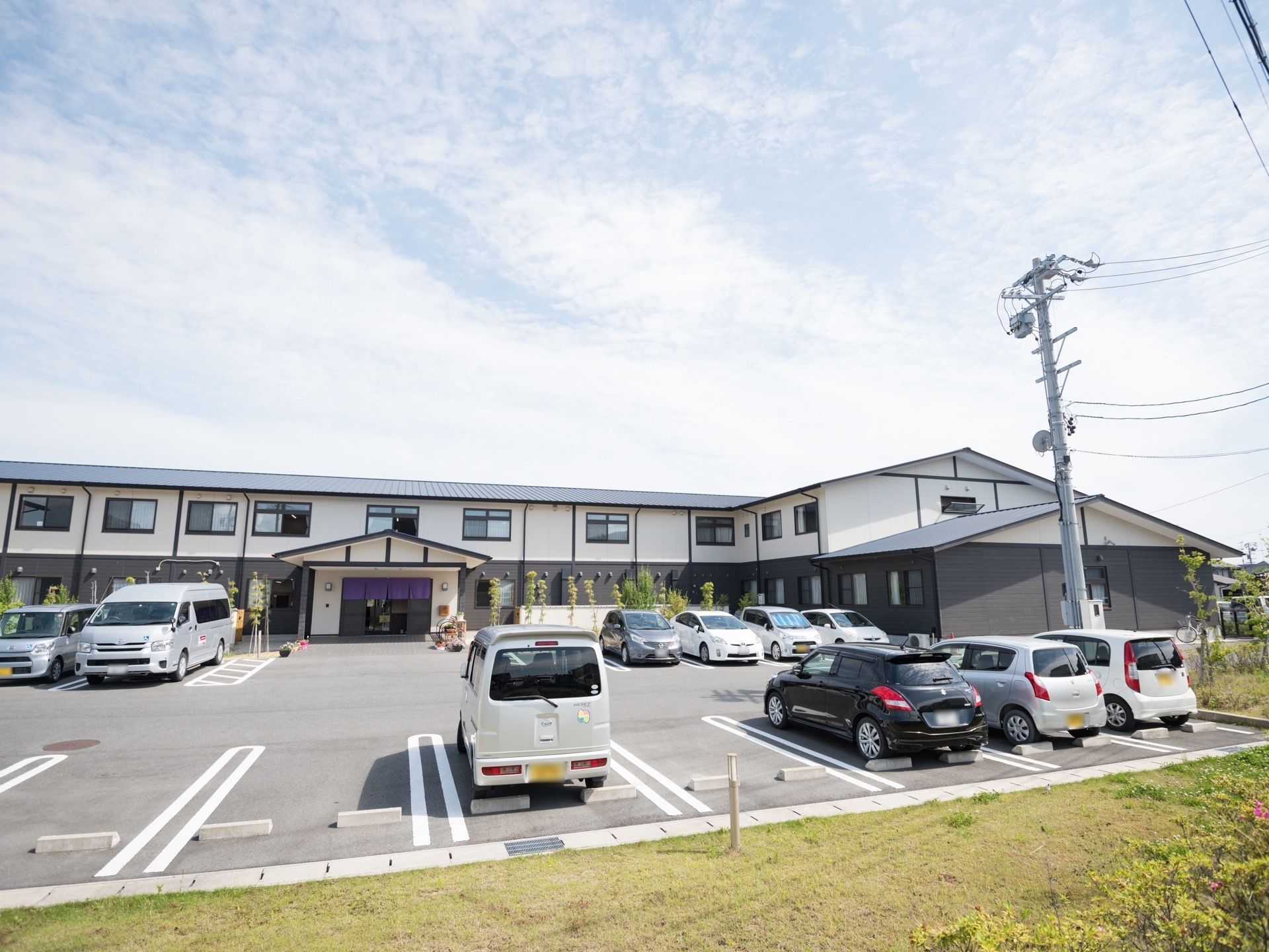 建物の前方には広いアスファルトの駐車場があり、車が多数停まっている。その手前には芝生が植えられたスペースがある。