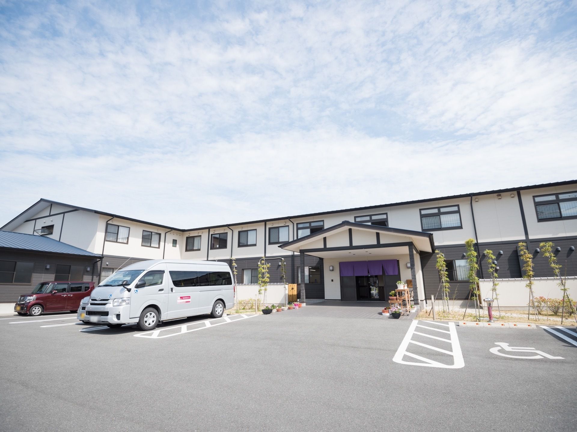 駐車場から建物へはフラットで段差なく移動できる。建物の周囲には植物が植えられていて、玄関付近に鉢植えの花が置かれている。