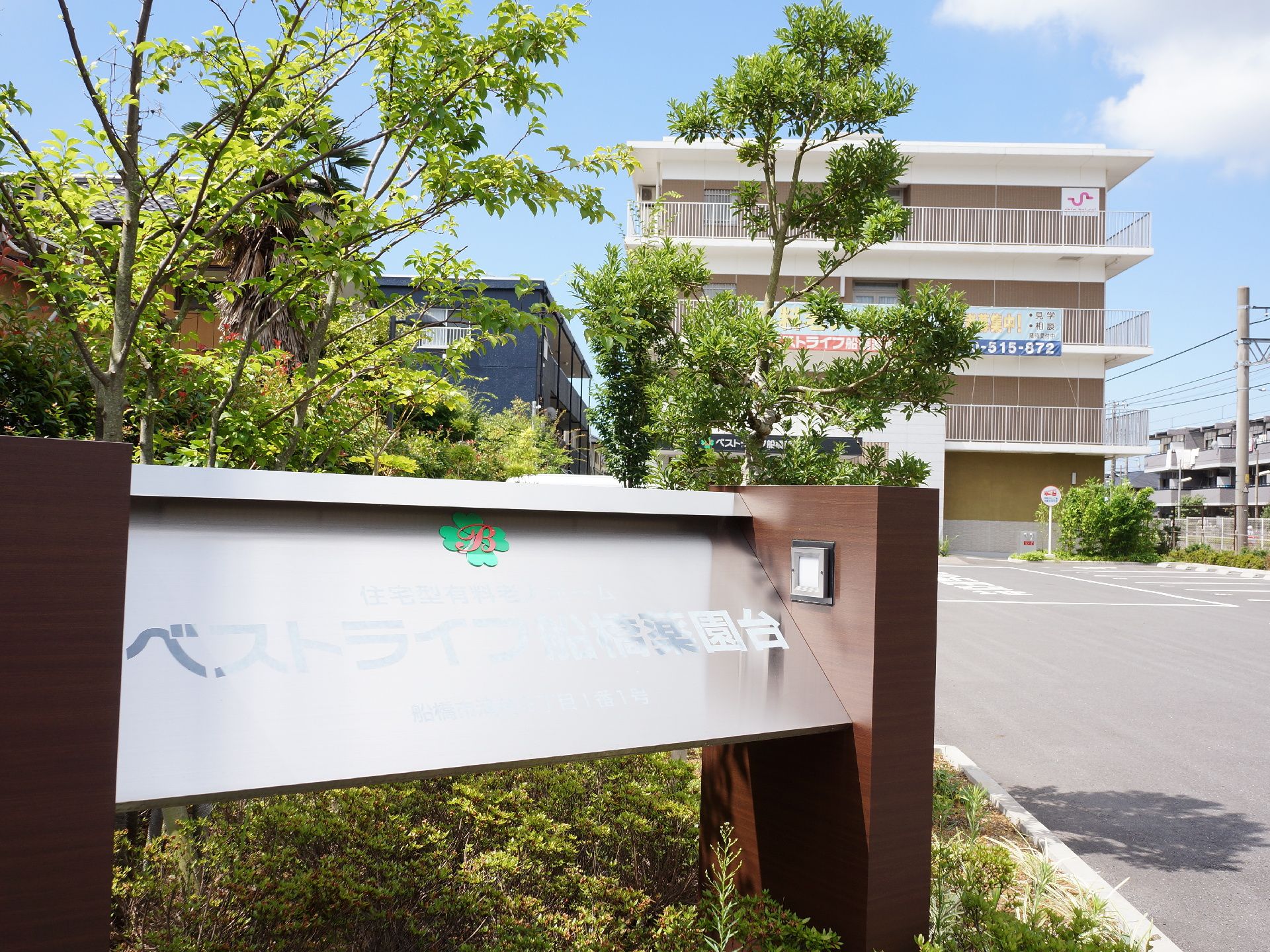 駐車場が完備されている。建物の周りには、綺麗に手入れされた木々が植わっている。施設名が書かれた看板がある。