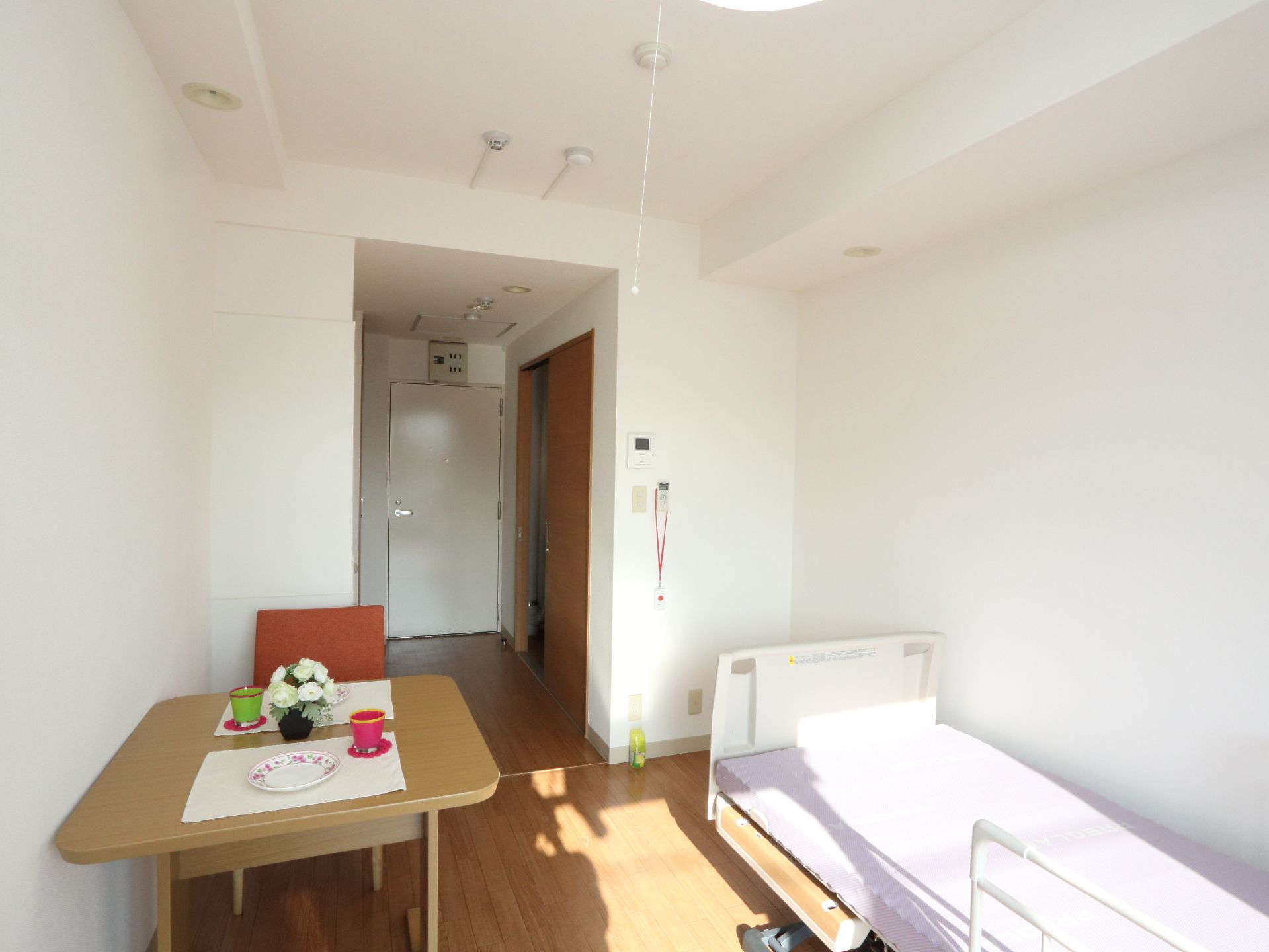 部屋にはテーブル、いす、ベッドなどがあります。日当たりがよく快適に過ごせます。壁には緊急用呼び出しボタンが設置されているので安心です。