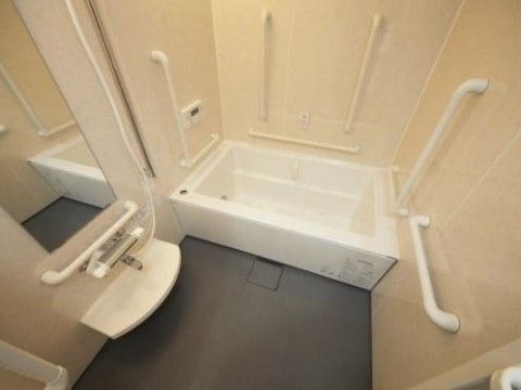 オフホワイトの壁とグレーの床の清潔感のある浴室。白い一人用の浴槽の周りには多くの手すりが設置されている。