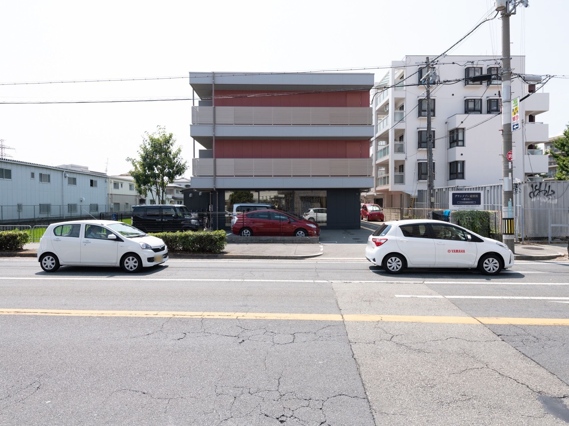 道路に面した、３階建ての施設である。施設名が記載されたプレートがある。駐車スペースがあり、数台の自動車が駐車している。