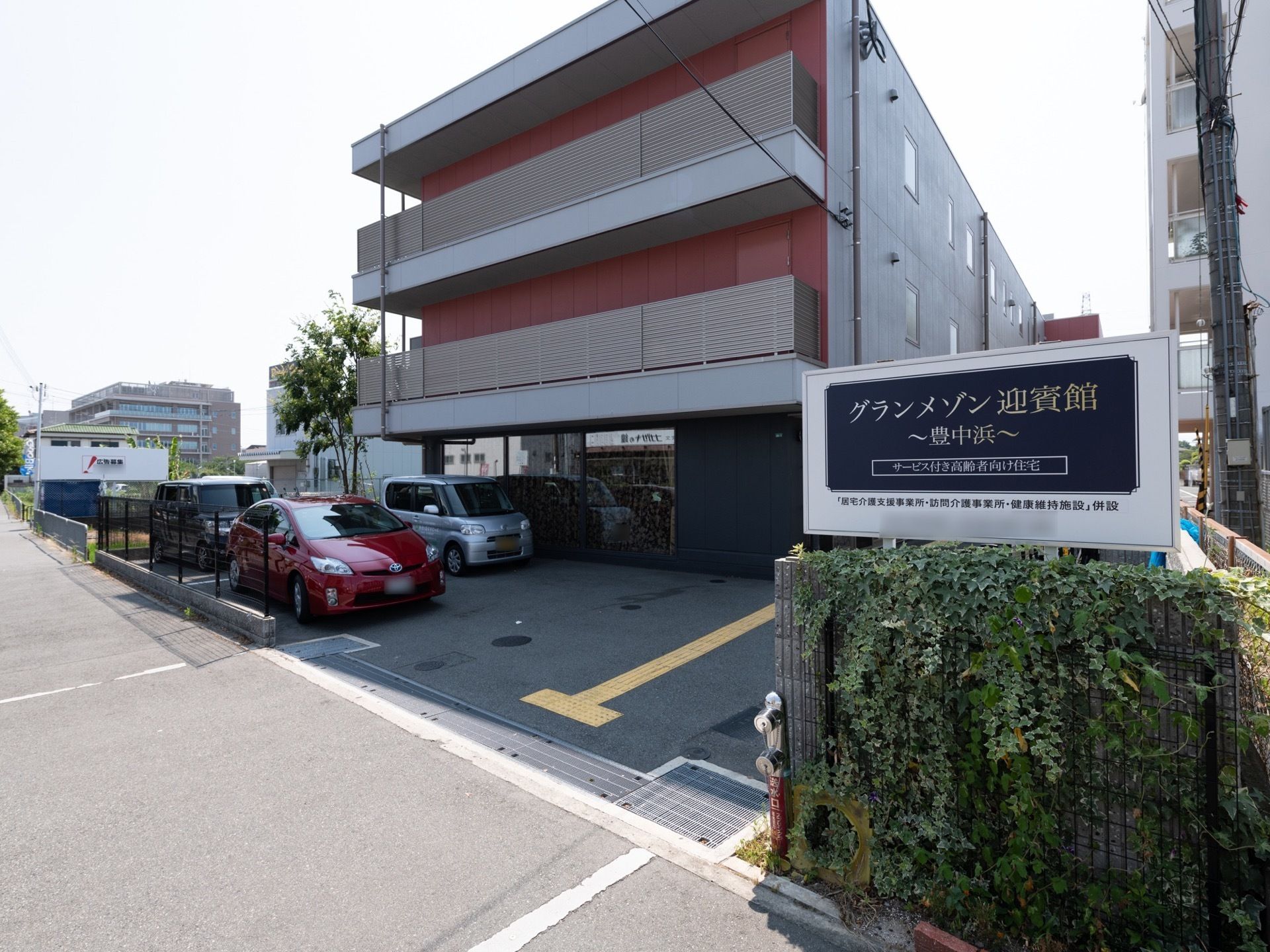 濃淡が異なるグレーを基調として、ベランダ内の赤い壁が目をひく外観である。道路側に駐車スペースを用意している。