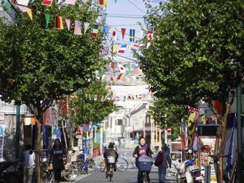 万国旗が飾られた商店街。青々とした街路樹や、店舗ののぼりが立ち並ぶ中を、自転車や徒歩の人々が行き交っている。