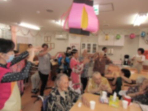 ピンク色の提灯のような飾りが天井に付けられ、テーブルに座った高齢者を囲んで複数の人が踊っている共用スペース