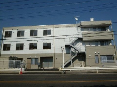 ホワイトとチャコールグレーの3階建ての建物。道路に面しており、ガードレールが設置されている。非常用の階段が見える。
