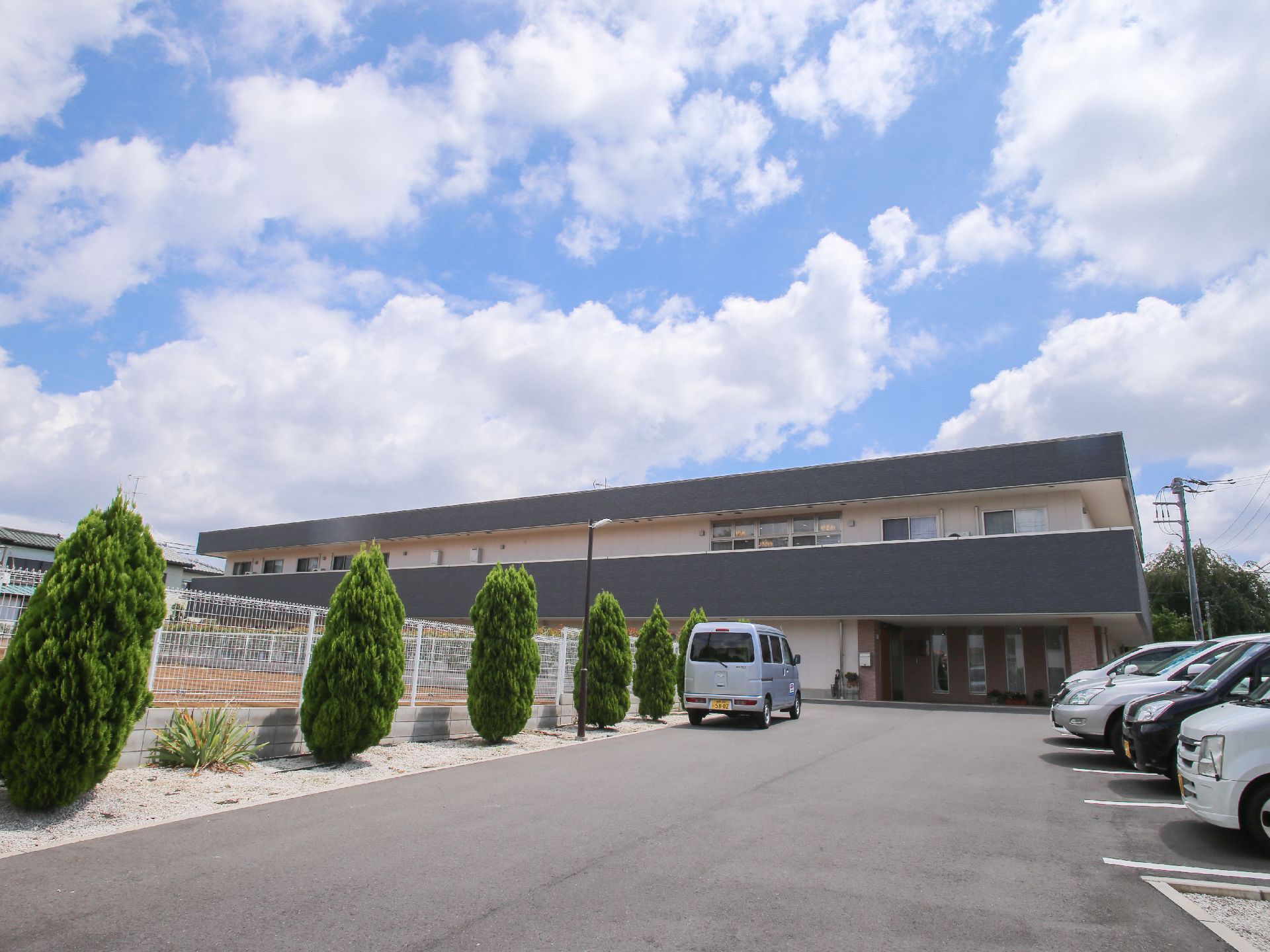 2階建ての建物の前は、広い駐車場になっている。敷地の境はフェンスで囲まれている。駐車場には木が植えられている。