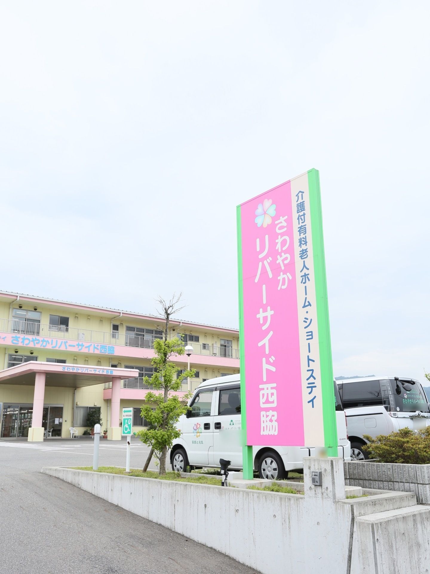 黄色とピンクが印象的な３階建ての建物であり、正面には施設名が大きく書かれている。施設の周りには広く駐車スペースがとられている。