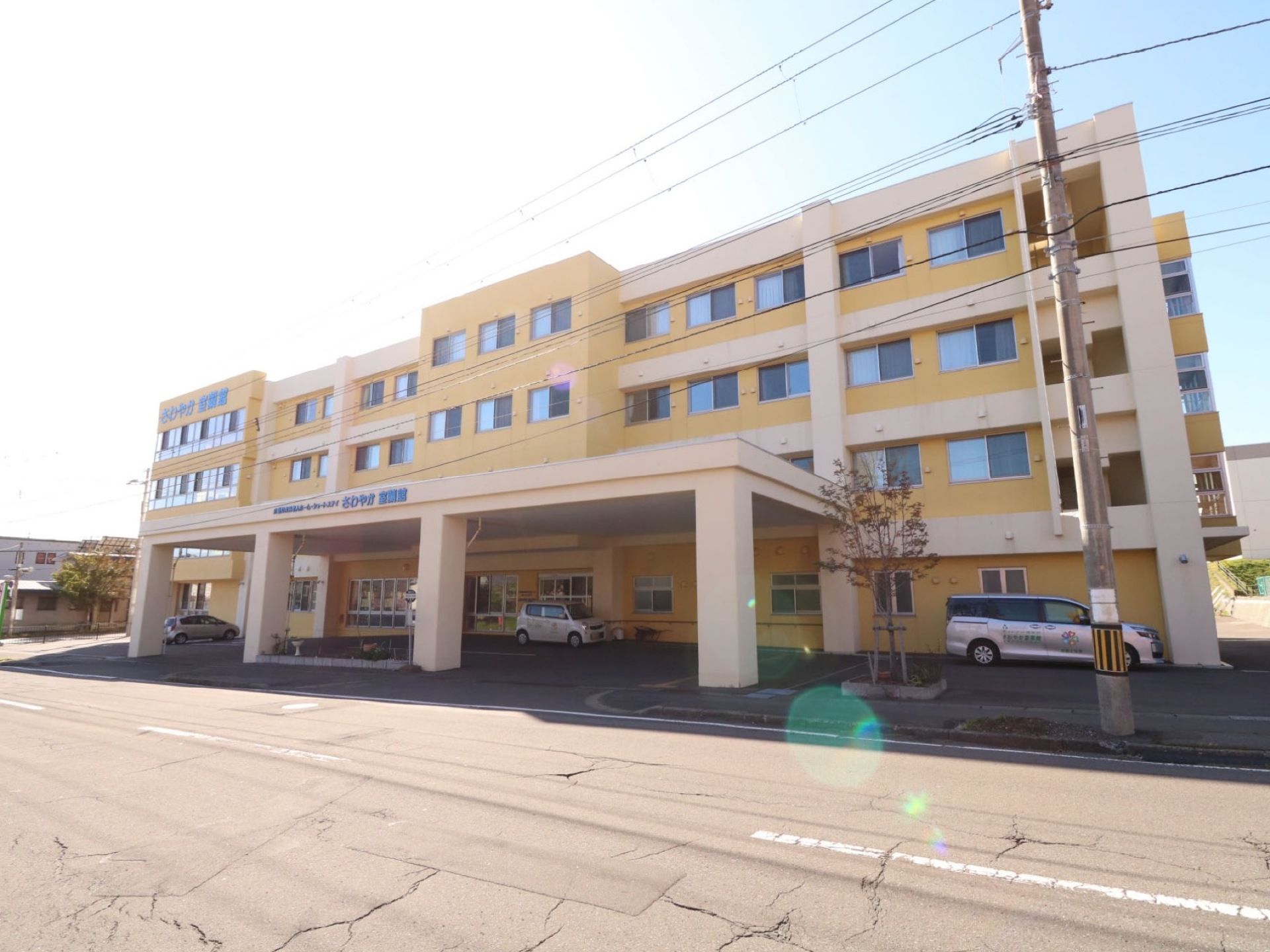 クリーム色の４階建ての建物が建っている。１階部分には施設名が表示された大きな雨よけがあり、車が停められている。