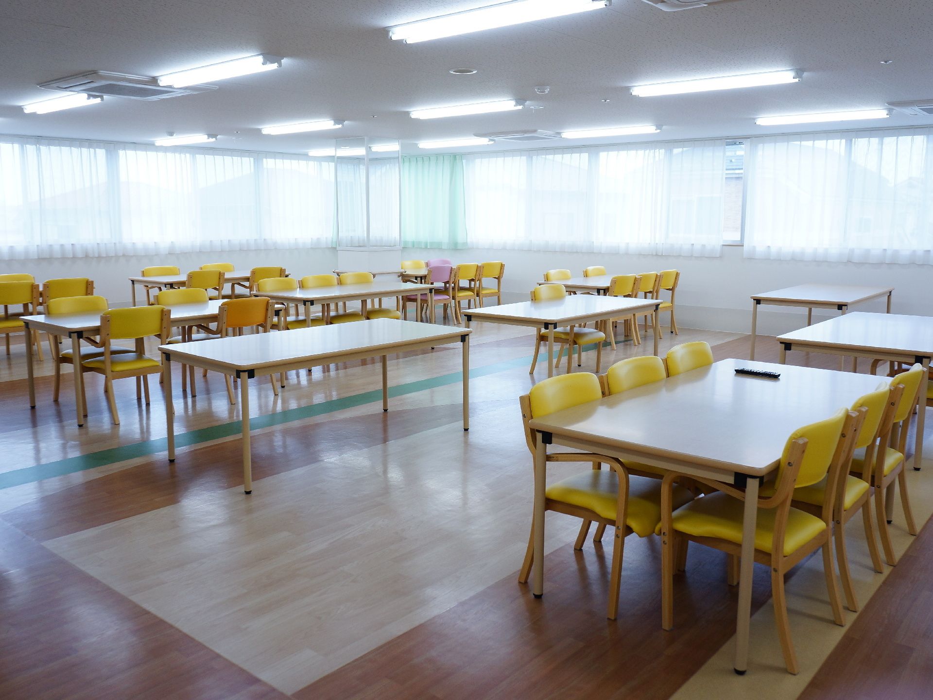 2面に大きく窓が取られた木目調のシート張りの床にテーブルが並んでいる食堂