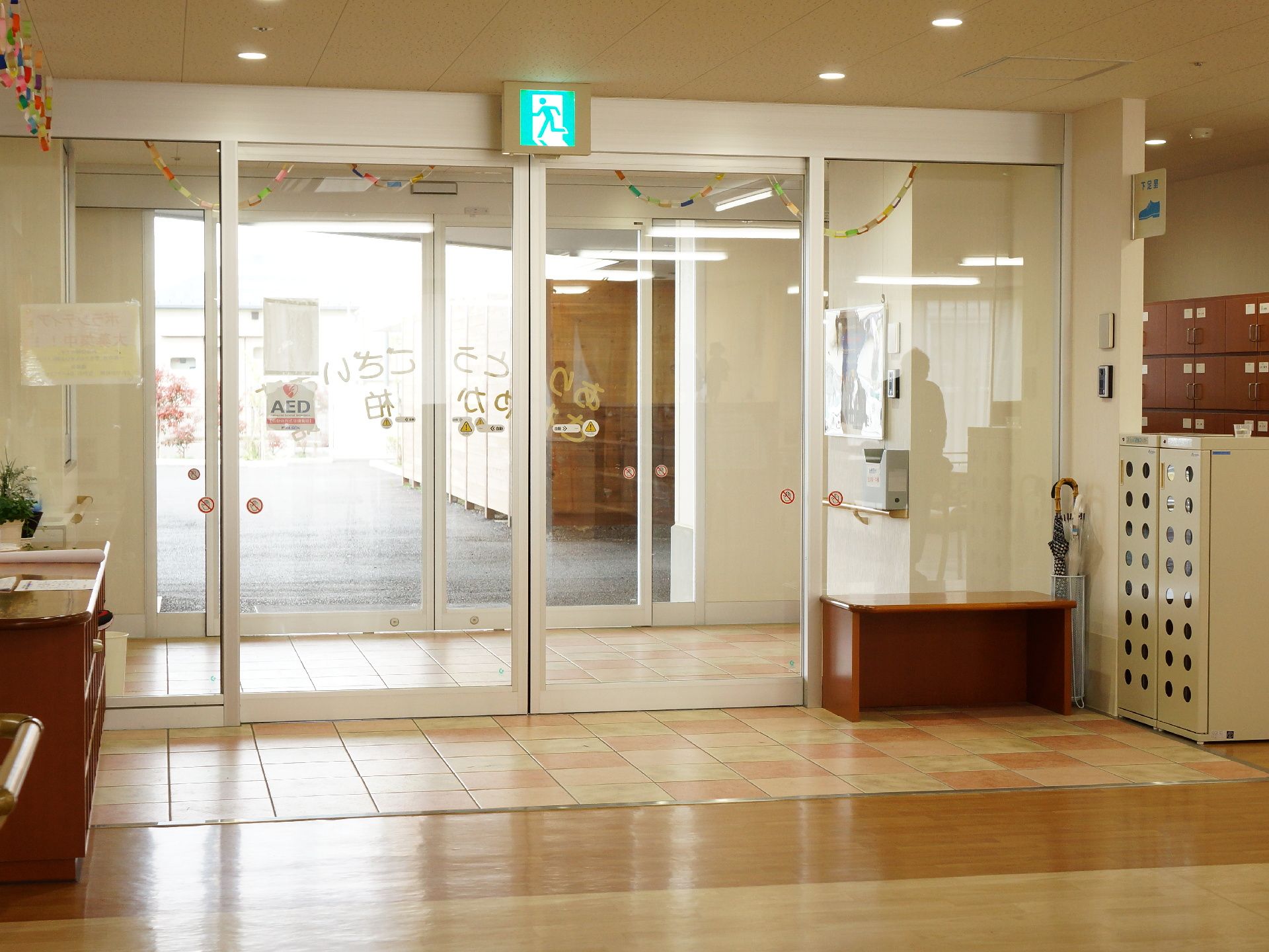 自動ドアの上に非常口の案内板がある。自動ドアの隣に扉付の大きな下駄箱がある。傘立てや腰掛も置かれている。