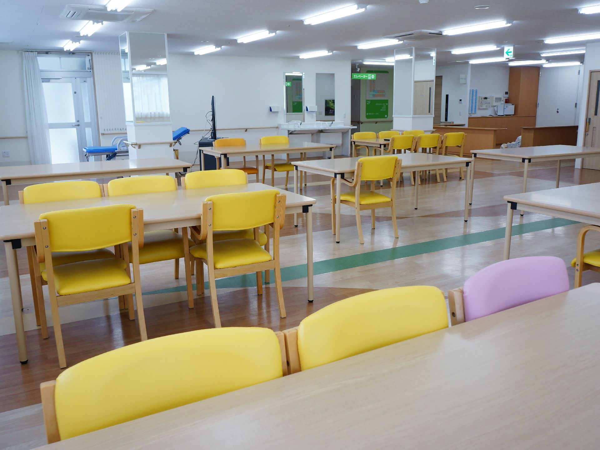木製のテーブルと黄色と薄紫の椅子が何個も置いてある。壁には手摺があり、洗面台が2個ある。ベッドとテレビも端にある。