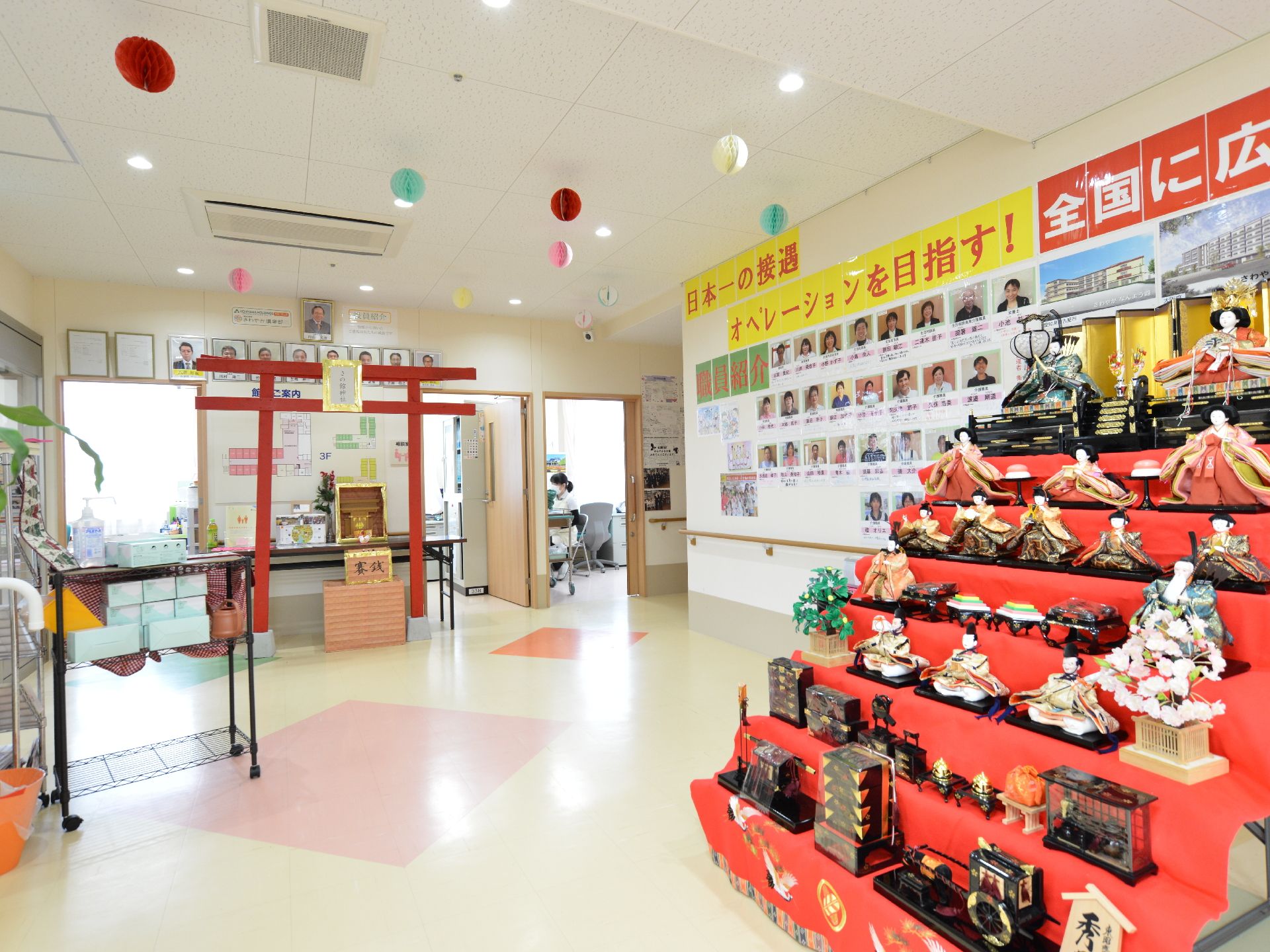 豪華な雛壇、鳥居に祠と賽銭箱が飾られている。壁一面にはスタッフの写真や会社の案内が貼られている。手摺もある。