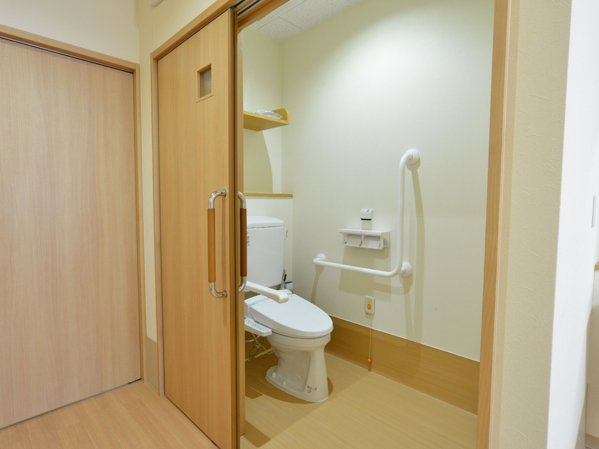 木製のスライド式の扉がついており中の壁にはL字型の手すりとナースコールが設置されており、手前に便座がある。