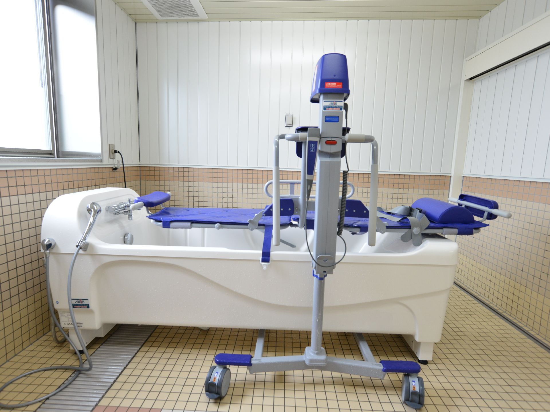 左側の窓から陽光が差し込んでおりタイル張りの部屋を照らしている。入浴のための介護専用器具が2台用意されている。
