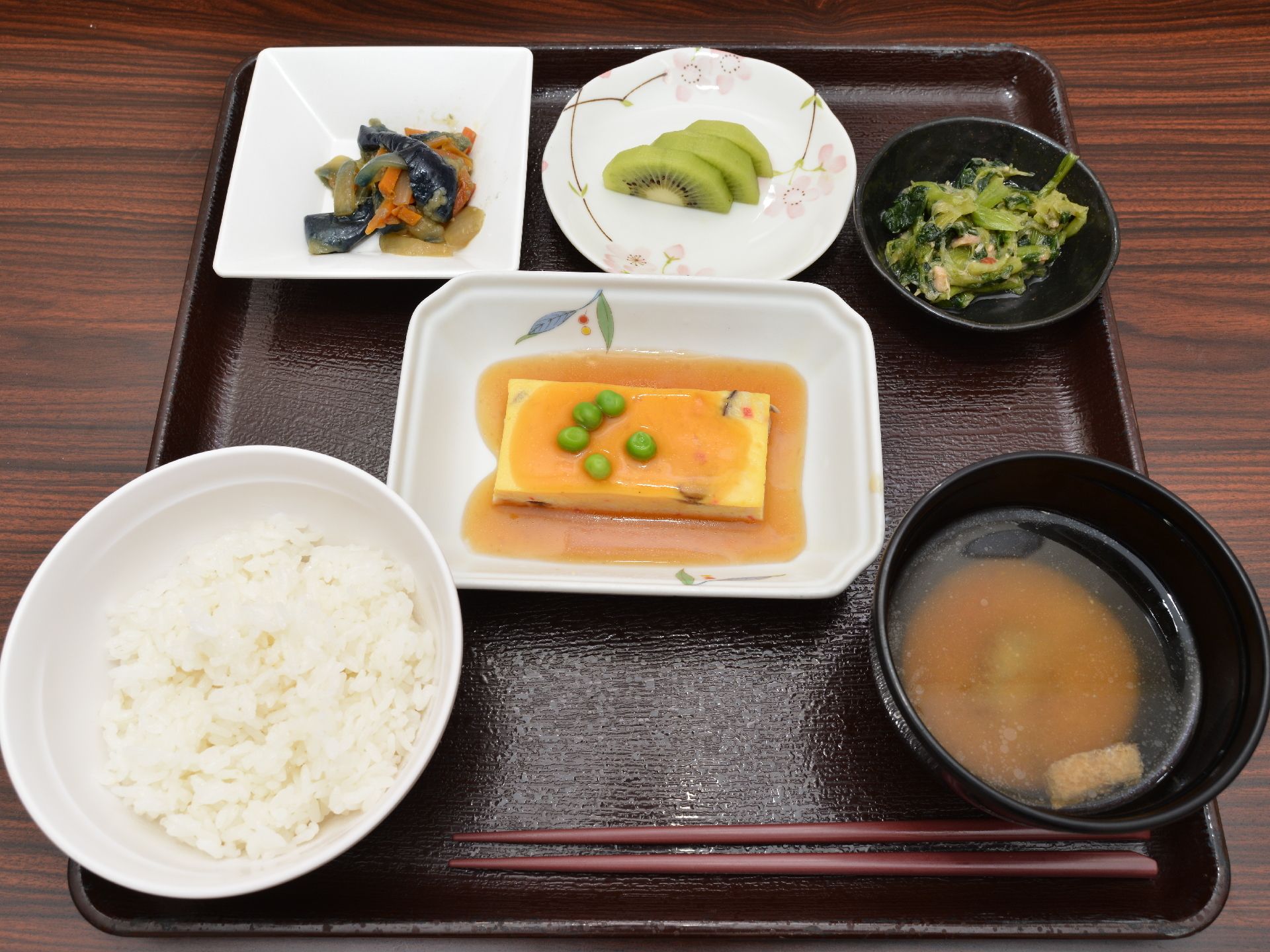 黒いトレイに栄養バランスが考えられた食事がのっている。あんかけのかかったメインと小鉢にデザートもついておりご飯と味噌汁ものっている。