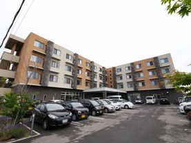 ４階建てのＬ字型の住宅がある。建物の前は舗装された駐車スペースになっていて、20台以上の車が停められている。