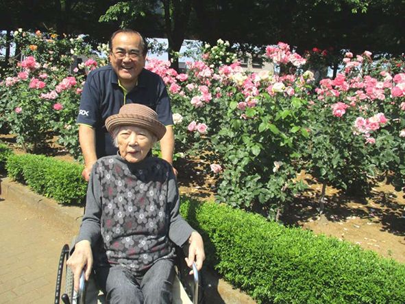 花の咲いた花壇の前で男性職員に車椅子を押してもらっている高齢女性