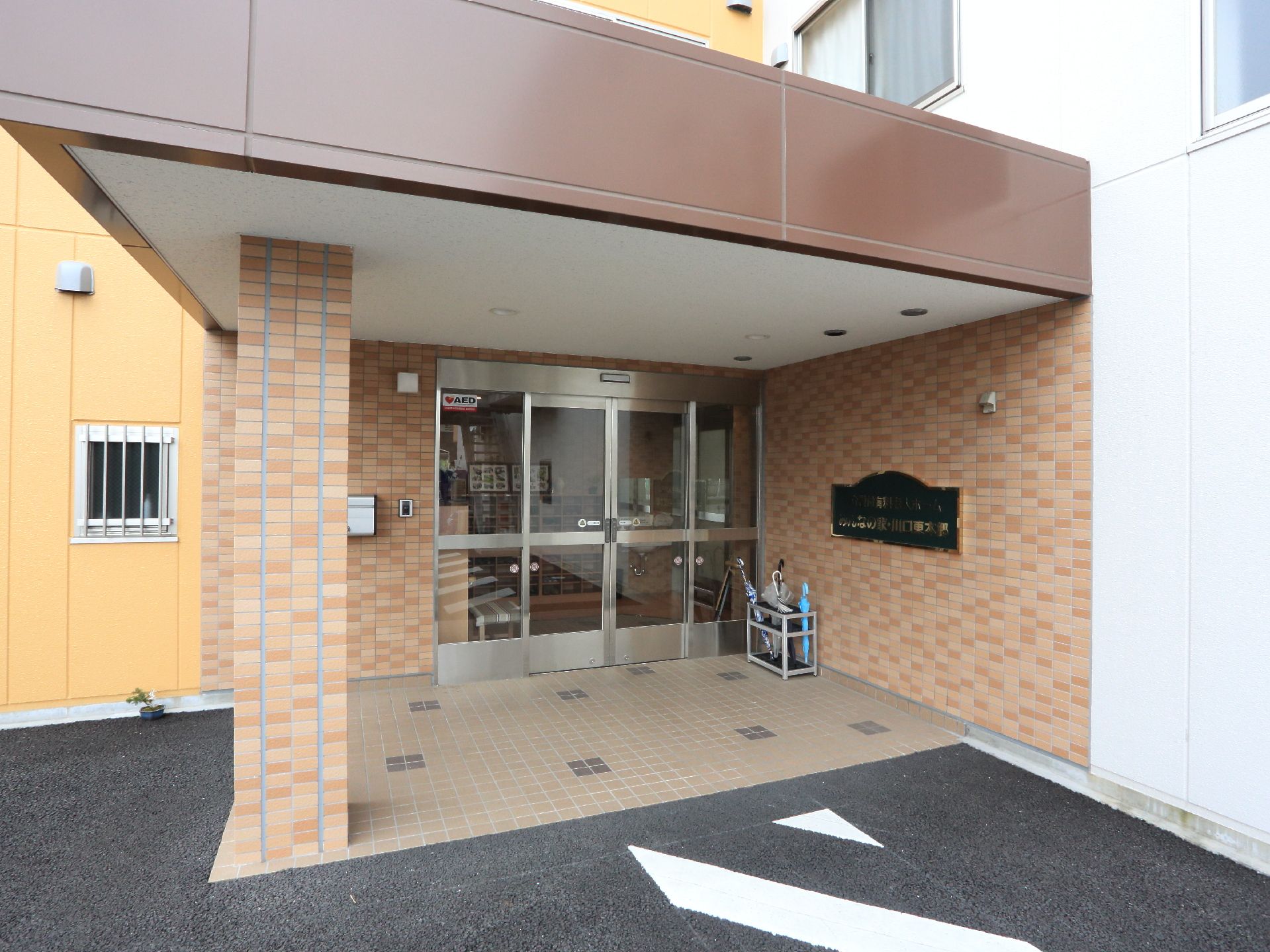 段差の無い入口。自動ドアの前には屋根が設置されている。壁には傘立てと施設名が書かれたプレートがある。