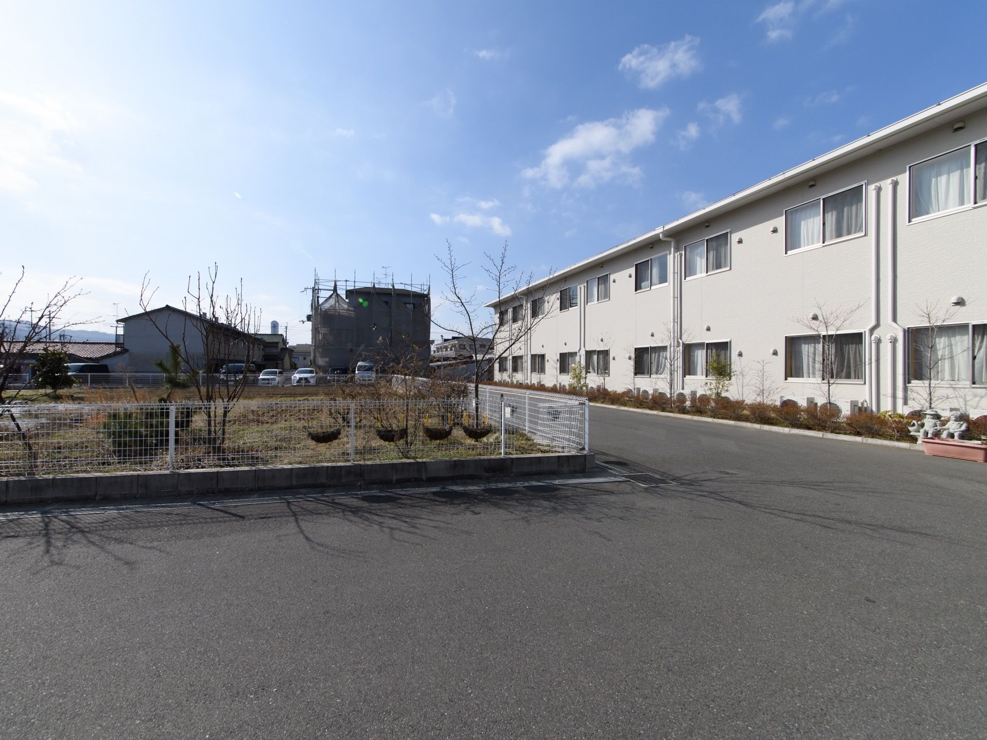 草木の生えた場所とフェンスによって隔てられたアスファルトの通路がある。通路に沿ってクリーム色の2階建ての施設がある。