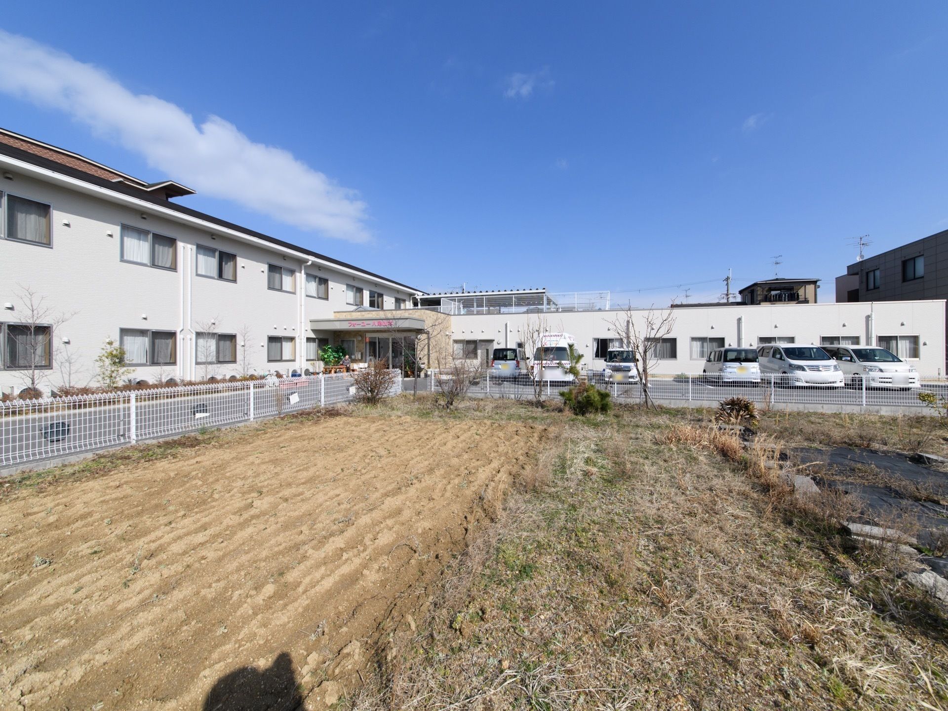 奥に施設の玄関と駐車場が見え、フェンスに囲まれた畑がある。施設の玄関とフェンスの間にはアスファルトの道がある。