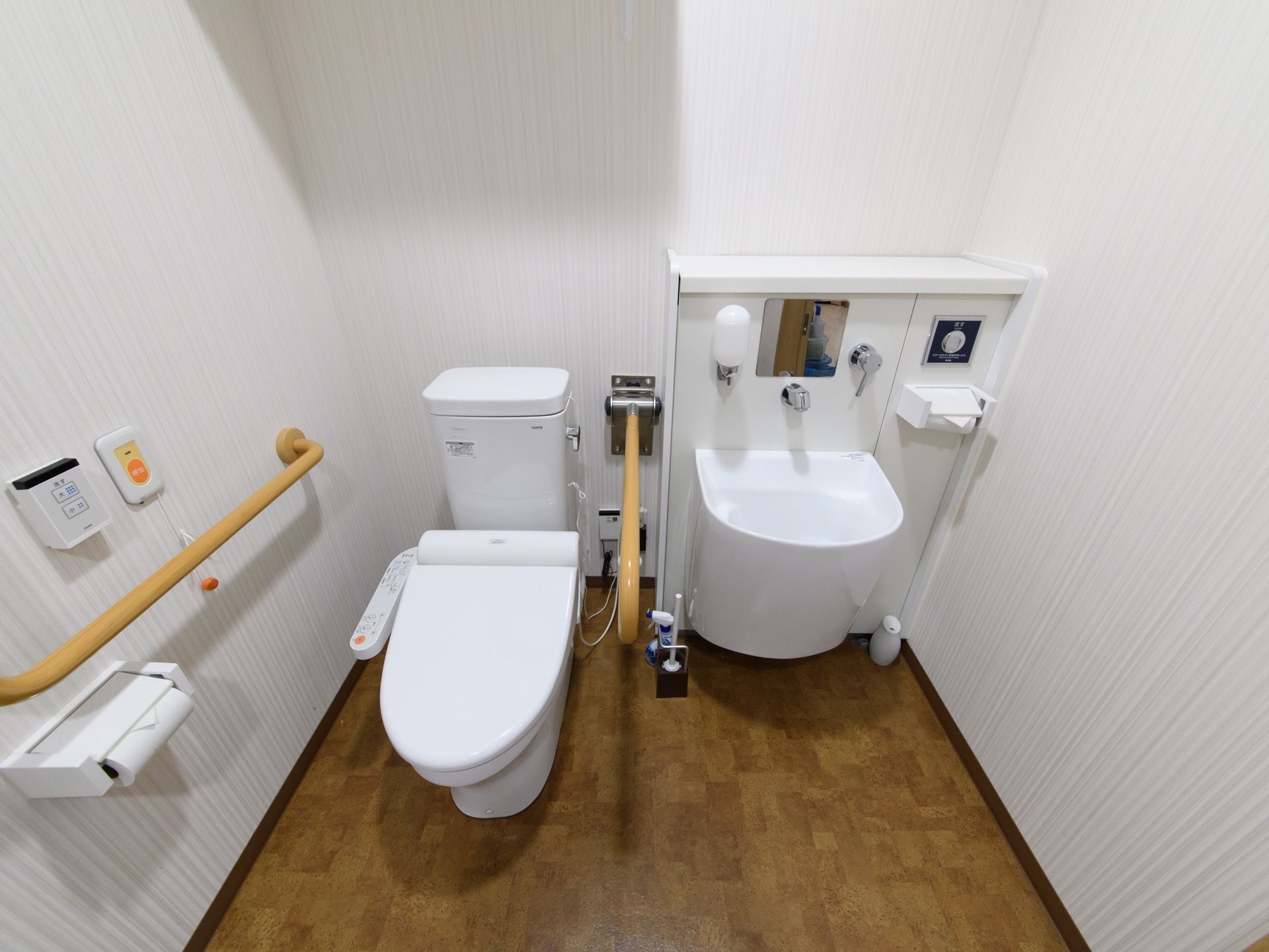 壁が白、床が木目調、便器の両横に手すりのついた手洗い場とセットになったトイレである。壁には緊急用の呼出ボタンが付いている。
