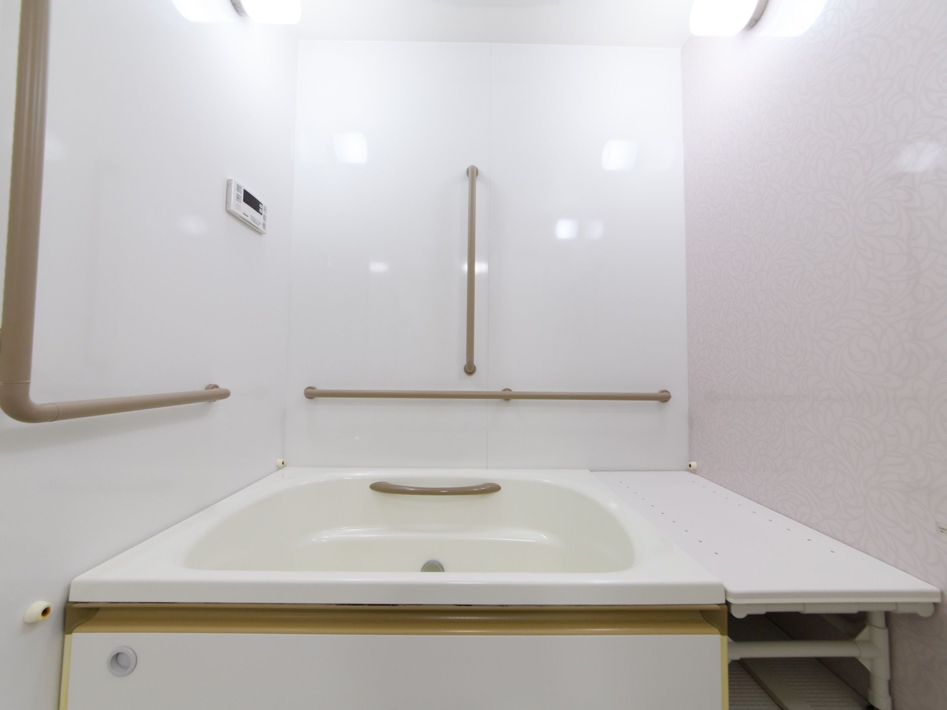 壁、天井、浴槽が白で構成されている。壁には白の照明器具と茶色の手すりが縦横の二方向の物が付いている。