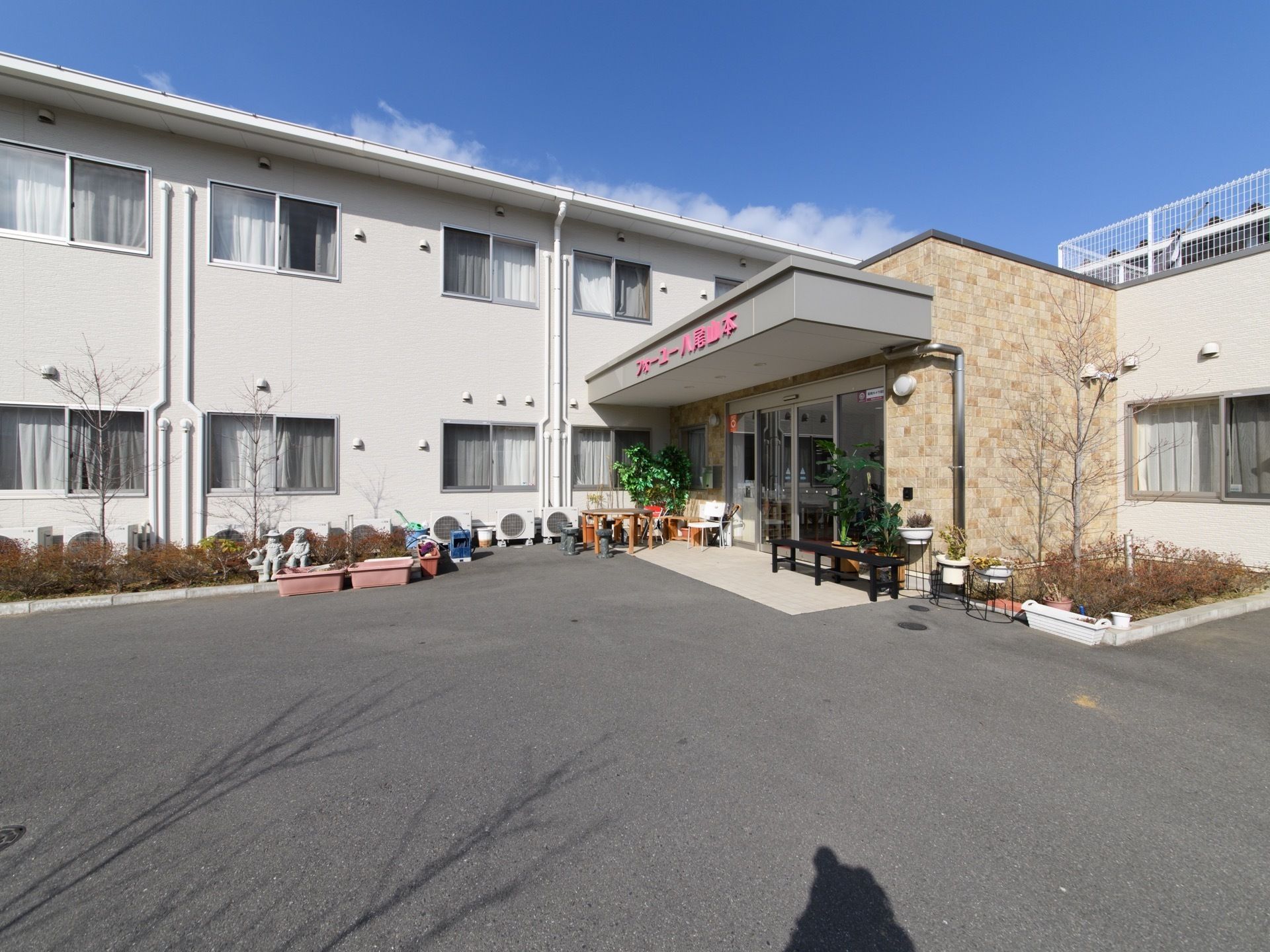 施設は２階建ての白い建物で、エントランスがあり、屋根の部分にはピンク色の文字で施設名が表示されている。自動ドアで施設に入ることができる。