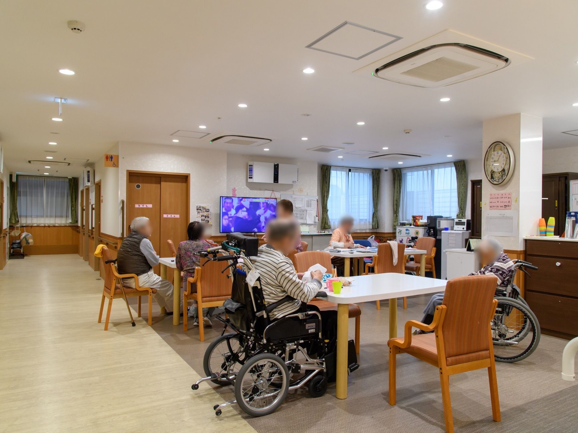 白いテーブルが３台あり、入居者様が思い思いに過ごしている。車椅子をご利用の方がいる。大きいテレビが置かれている。