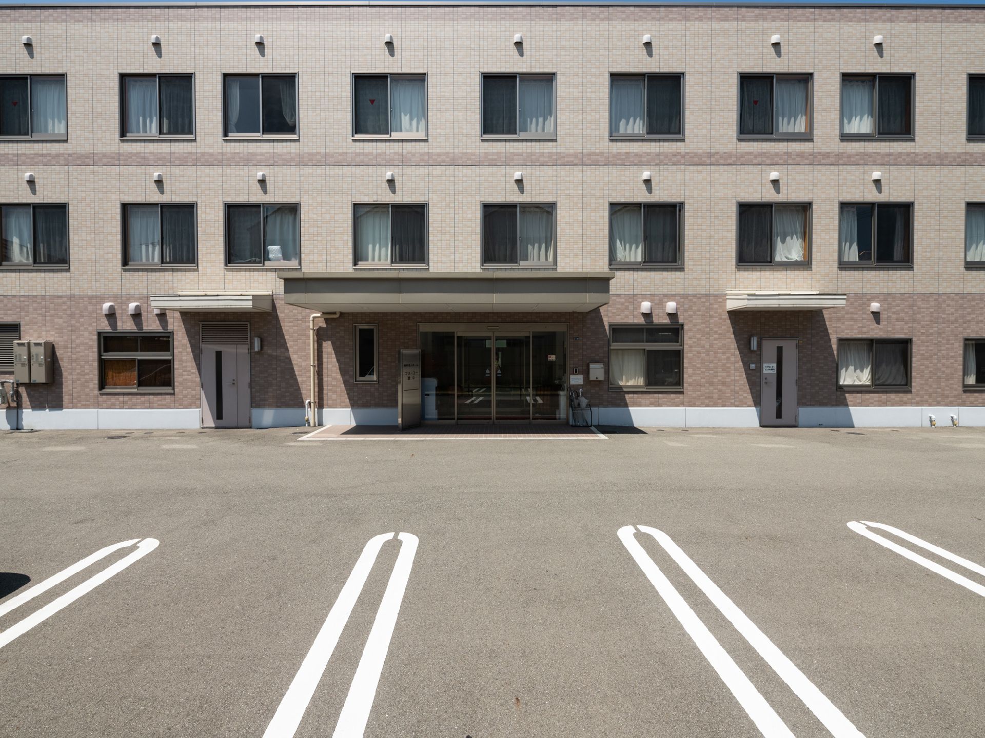 ３階建ての新しい建物。外壁は淡いベージュと茶色で凹凸がない。敷地内は舗装されており、駐車場がある。玄関前にはひさしがある。