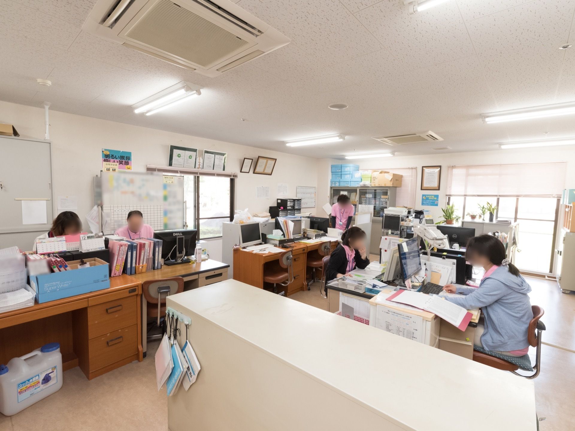 ピンク色の制服を着た５人の職員がそれぞれに仕事をしている。机と椅子が向かい合わせに並び、机の上にはパソコンが置かれている。