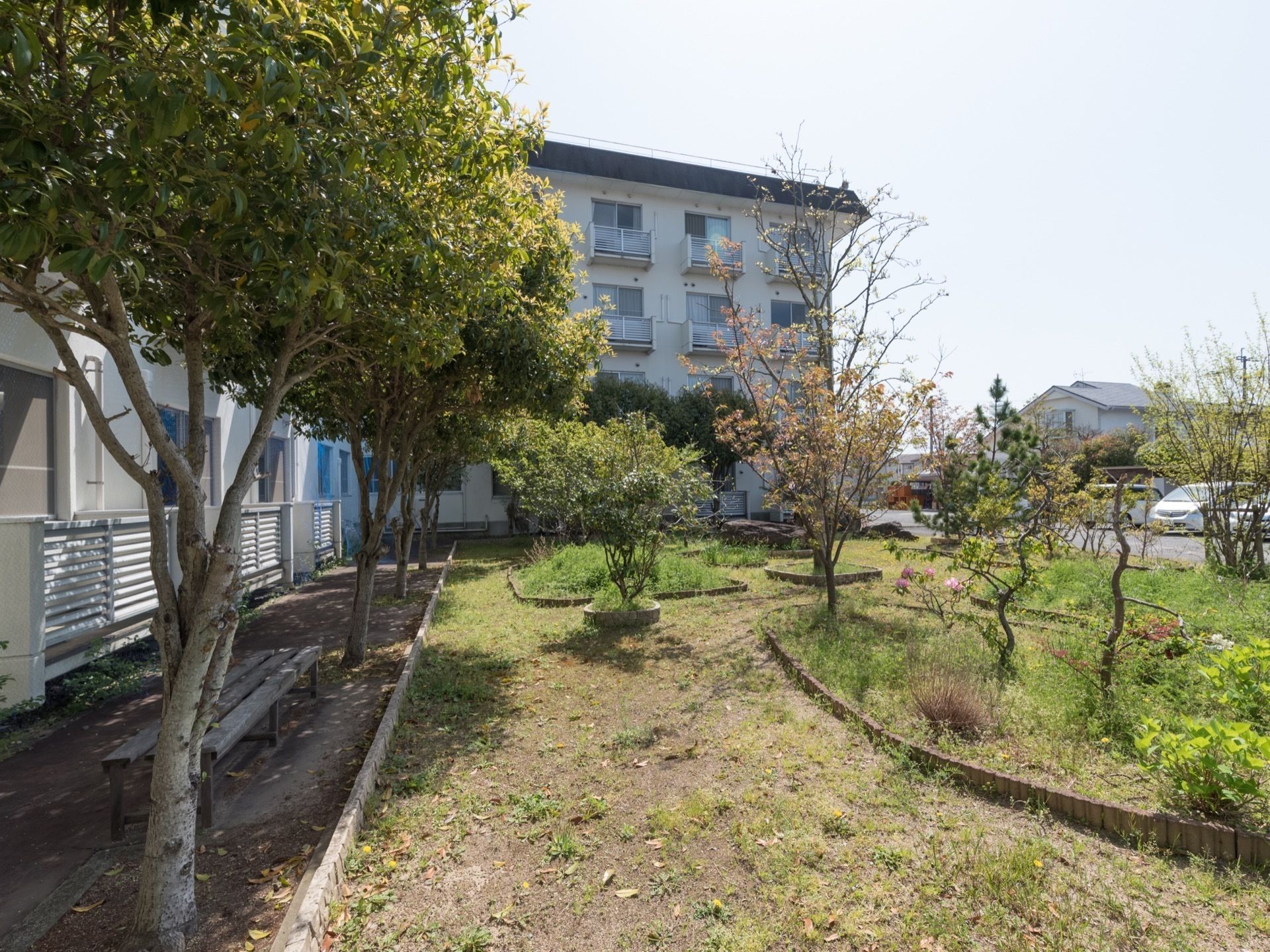 広い敷地にはたくさんの樹木が植えられている。木陰にベンチが１台置かれている。庭の前は駐車場になっていて、車が数台停まっている。