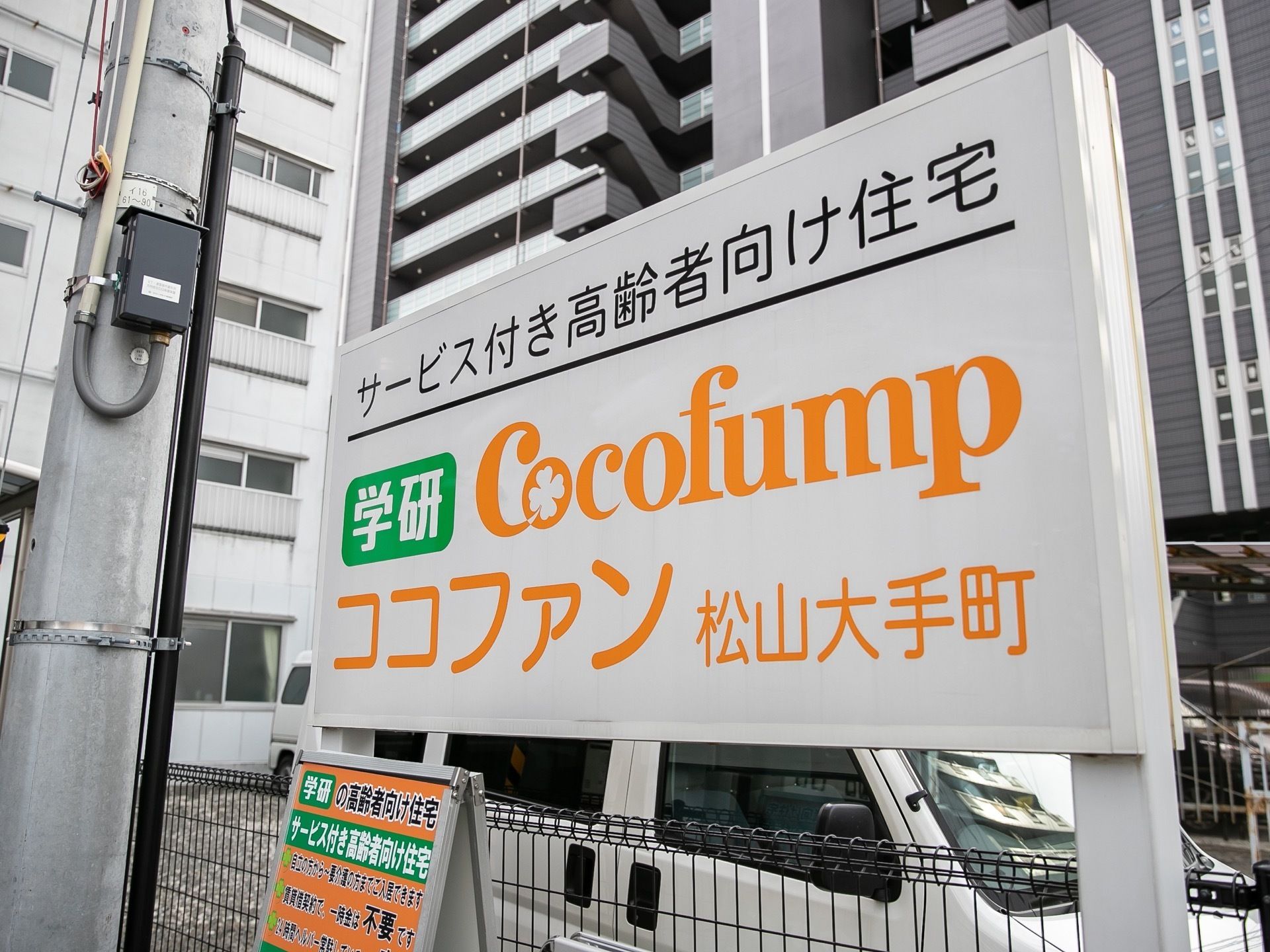 黒色のフェンスの前には施設の看板が立てかけられている。看板は白色で文字は黒色とオレンジ色、緑色が使われている。横には電柱がある。