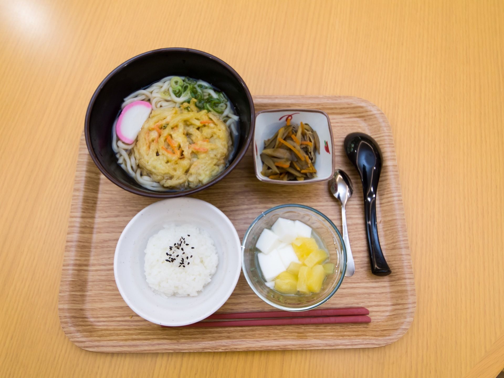 茶系の模様入りのトレイに、てんぷらとかまぼ、ネギがのったメインのうどんとご飯、小鉢、フルーツをのせている。お箸とスプーンなどを添えている。