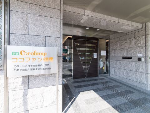 左に看板、奥にブラウンの自動ドアが見えるグレーの石造り風のエントランス
