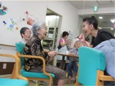 椅子に腰かけた高齢女性に向かってこぶしを振って笑顔で話しけている女性