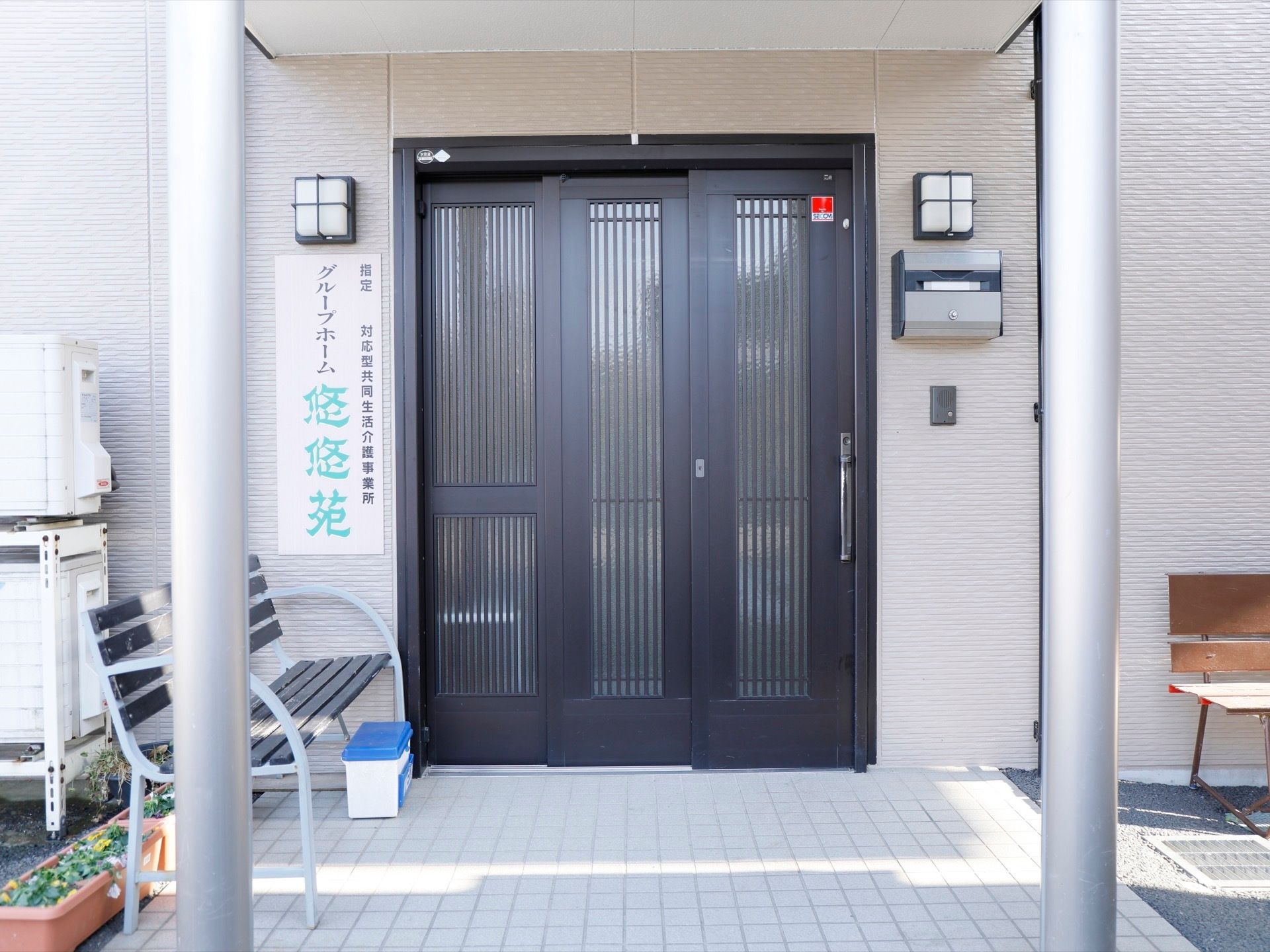 施設の玄関ドアは不透明ガラスをはめ込んだ引き戸である。前面の床はタイル敷きで、休憩用のベンチを用意している。