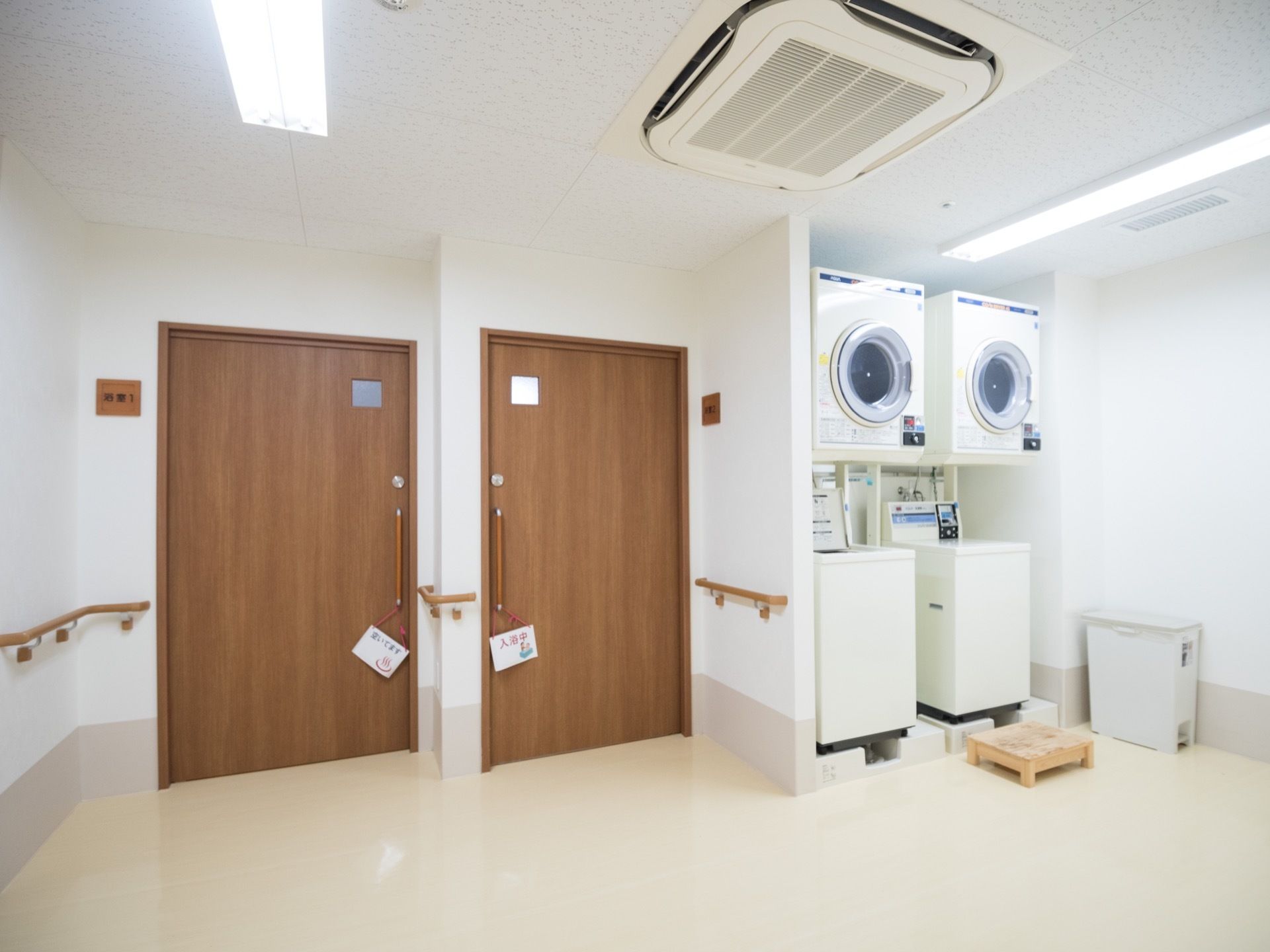 白を基調とした清潔感あふれるランドリー室。洗濯機と乾燥機が２台ずつ設置されており、横には浴室へと続く扉がある。