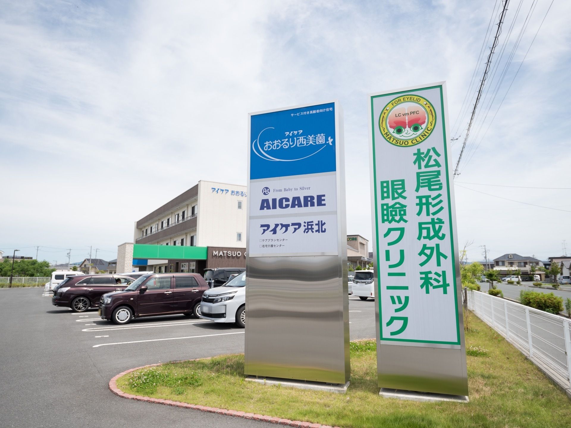 静かな環境の中にあるホームで、敷地内の駐車場に数多くの車が停車している。施設入口には、大きな看板が２つ設置されている。