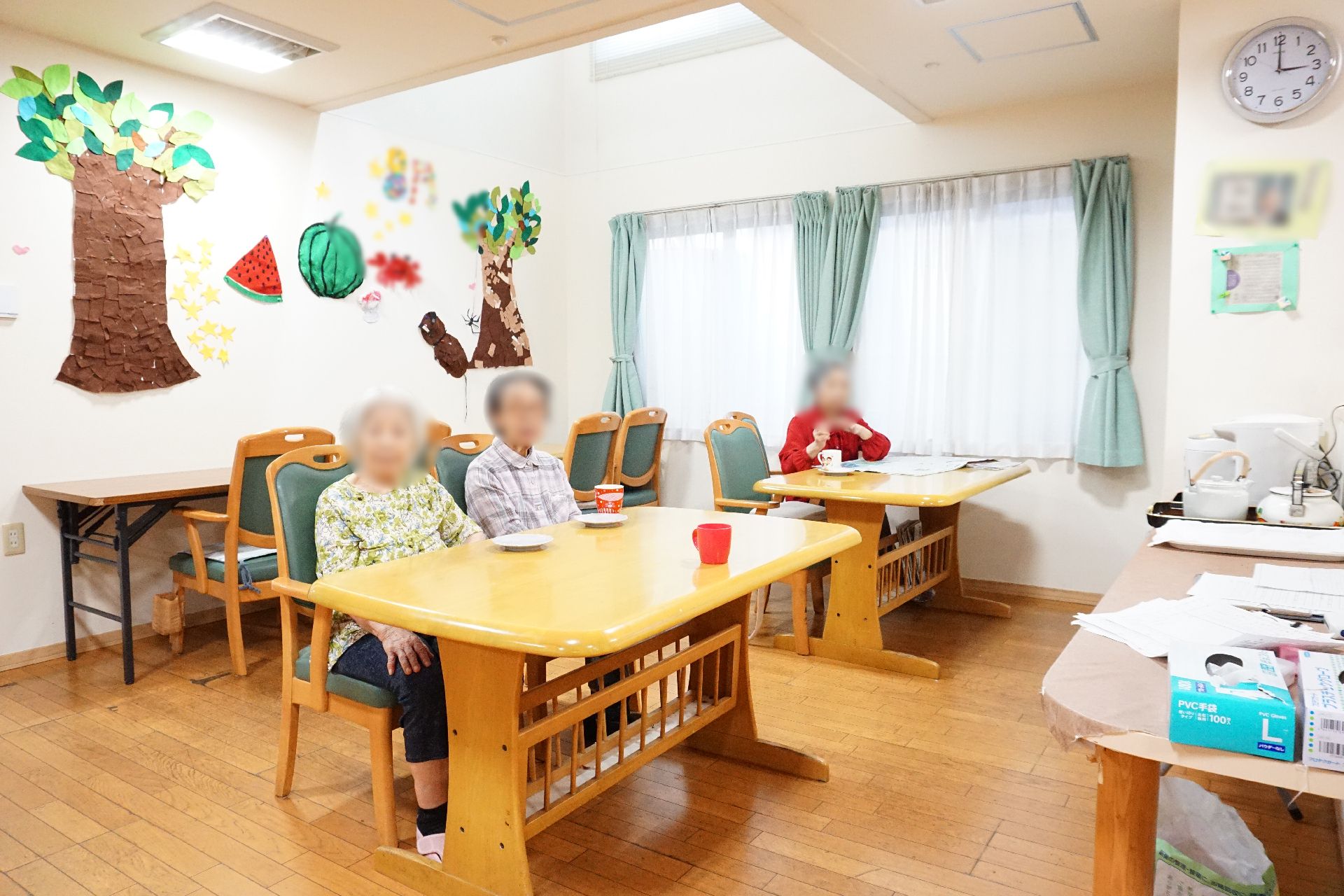 壁に手作りの展示物が飾られ、入居者がテーブルの前に座っているリビング